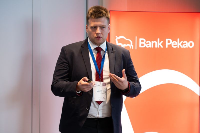 Zdjęcie przedstawia osobę w garniturze na szaro-czerwonym tle z logiem Bank Pekao.