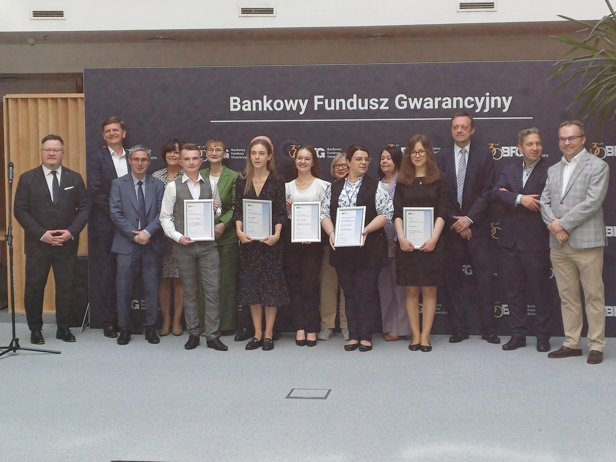 Grupowe zdjęcie uczestników i organizatorów wydarzenia z okazji wręczenia nagród lub certyfikatów przez Bankowy Fundusz Gwarancyjny. W pierwszym rzędzie stoi sześć osób trzymających dyplomy, za nimi grupa przedstawicieli w formalnych strojach. W tle ścianka z logo Bankowego Funduszu Gwarancyjnego.