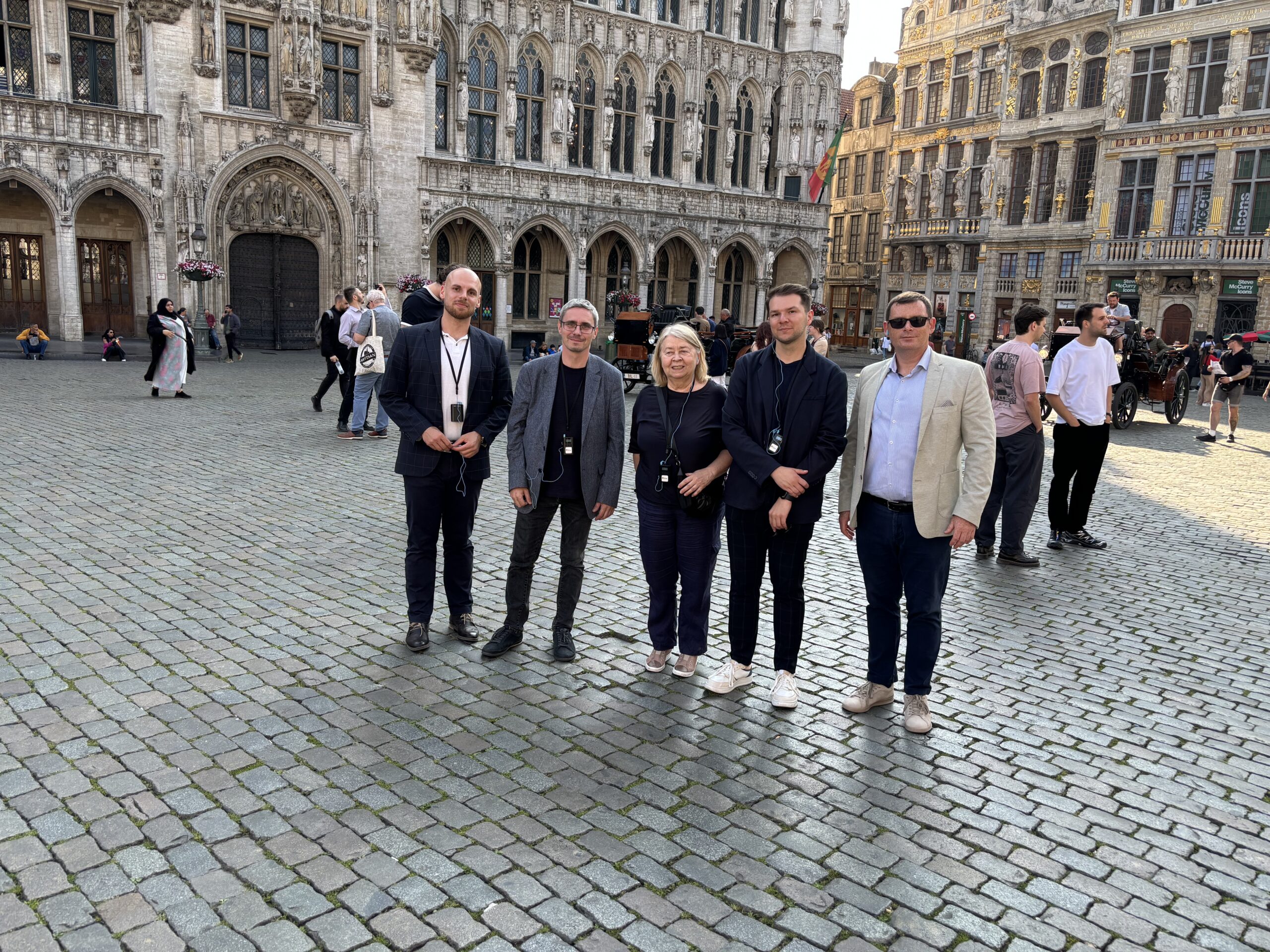 Na zdjęciu widocznych jest pięć osób stojących na wybrukowanym placu w zabytkowym centrum Brukseli. To pracownicy Katedry oraz przedstawiciele Santander Bank Polska, uchwyceni podczas zwiedzania. W tle znajduje się imponujący, historyczny budynek o bogato zdobionej fasadzie, z łukowatymi oknami i rzeźbami. Wokół poruszają się inni przechodnie, a słoneczna pogoda dodaje scenie turystycznego i przyjaznego charakteru.