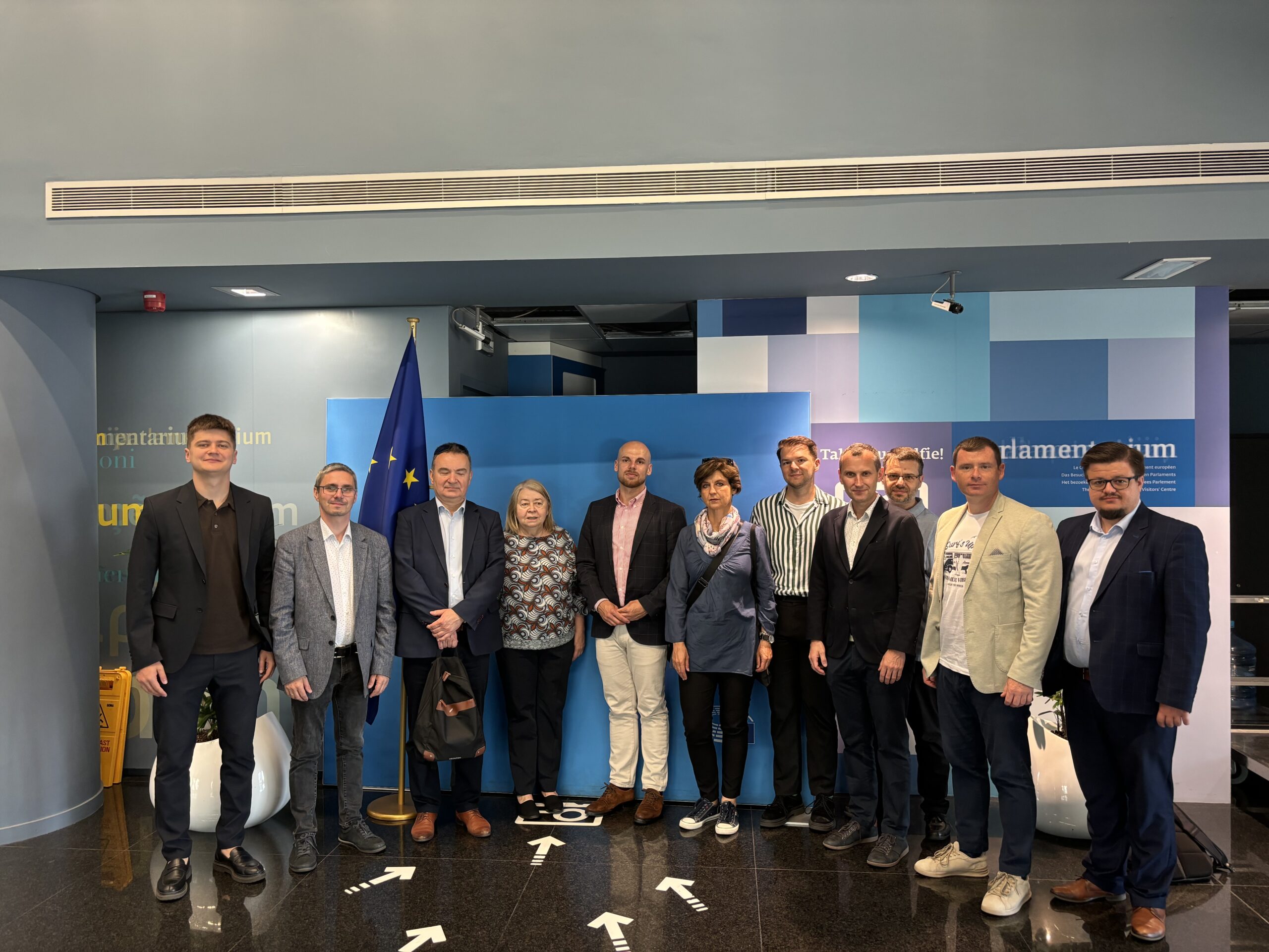 Na zdjęciu znajduje się grupa jedenastu osób stojących wewnątrz budynku, na tle ścianki z flagą Unii Europejskiej. To pracownicy Katedry oraz przedstawiciele Santander Bank Polska, pozujący do zdjęcia w Parlamentarium – centrum dla odwiedzających Parlament Europejski. Wszyscy są elegancko ubrani, część osób trzyma ręce w kieszeniach lub splecione przed sobą. W tle widoczne są napisy oraz elementy dekoracyjne związane z instytucjami europejskimi. Cała scena ma miejsce w nowoczesnym wnętrzu z marmurową podłogą.