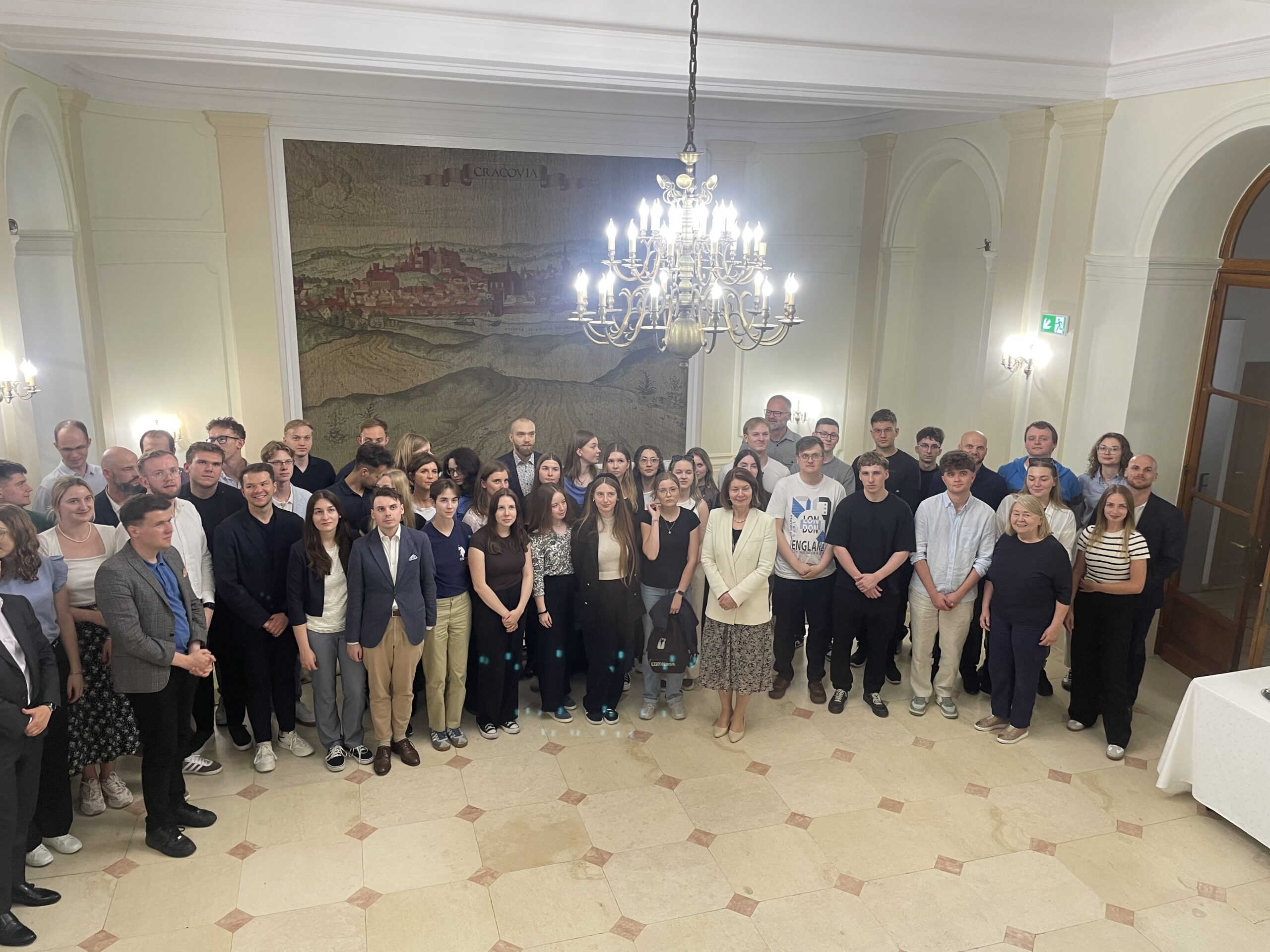 Zdjęcie grupowe wykonane we wnętrzu Ambasady RP w Brukseli podczas spotkania uczestników wyjazdu studyjnego z Panią Chargé d’Affaires Katarzyną Skórzyńską, stojącą pośrodku grupy. Uczestnicy pozują wspólnie w eleganckiej sali z żyrandolami i dużą dekoracją ścienną przedstawiającą panoramę Krakowa.