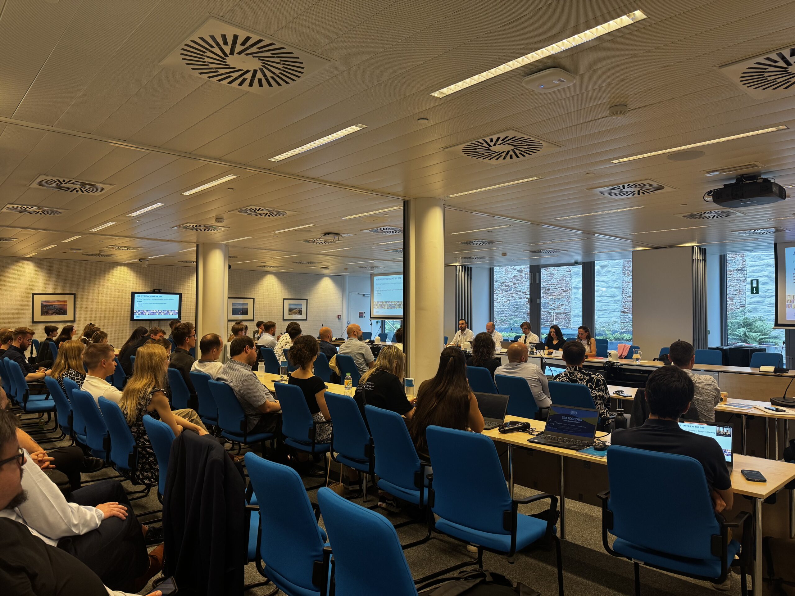 Na zdjęciu widoczna jest sala konferencyjna, w której odbywa się spotkanie uczestników wyjazdu studyjnego do Brukseli z ekspertami ds. restrukturyzacji i uporządkowanej likwidacji banku z Single Resolution Board (SRB). W pomieszczeniu znajduje się wiele osób siedzących na niebieskich krzesłach przy stołach. Przy stole prezydialnym na końcu sali zasiada kilka osób, pełniących rolę prelegentów lub prowadzących spotkanie. Na ścianach wiszą obrazy, a w sali znajdują się ekrany wyświetlające prezentacje. Atmosfera wydarzenia jest formalna i profesjonalna.