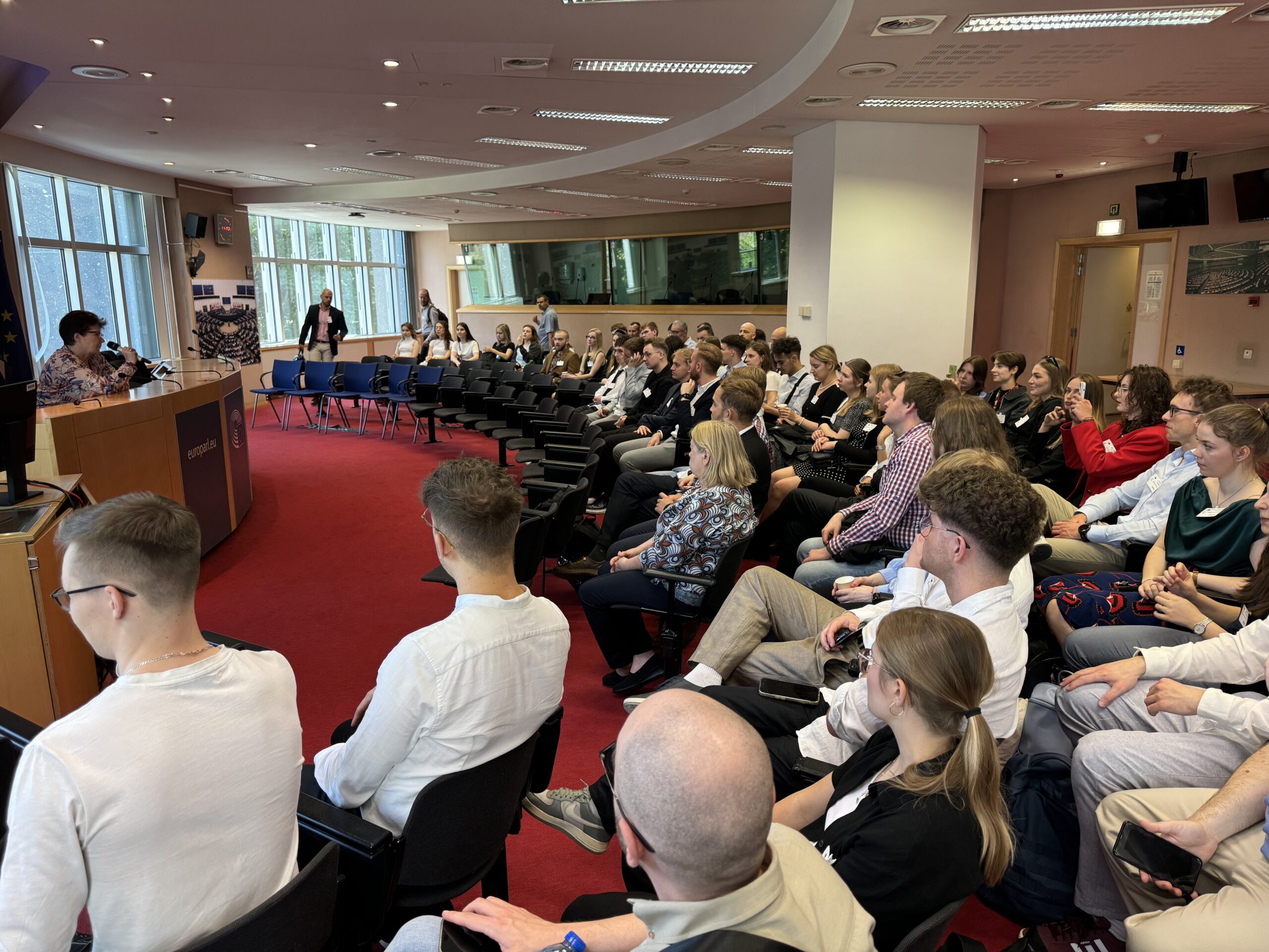 Na zdjęciu widoczna jest sala konferencyjna, w której uczestnicy wyjazdu studyjnego do Brukseli siedzą na krzesłach, zwróceni w stronę mównicy. Wykład prowadzi Pani Prof. Hanna Gronkiewicz-Waltz i dotyczy roli oraz zadań Rady do Spraw Gospodarczych i Finansowych (ECOFIN). Sala jest przestronna, z dużymi oknami po lewej stronie i czerwonym dywanem na podłodze. Część osób słucha uważnie, inne dokumentują wydarzenie za pomocą telefonów.