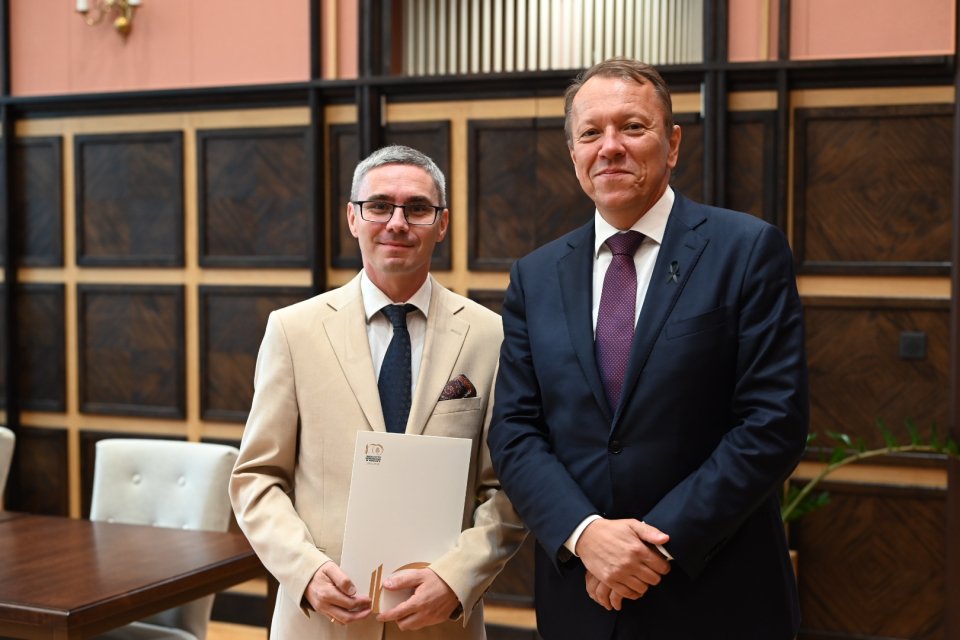 Na pierwszym planie znajduje się dwóch mężczyzn w formalnych strojach. Jeden z nich trzyma dokument w dłoni. Tło to eleganckie wnętrze, część budynku publicznego z drewnianymi panelami na ścianach i nowoczesnymi elementami wystroju. Dwóch mężczyzn są dobrze ubrani, co wskazuje na formalną okazję. Mężczyźni stoją obok siebie i patrzą w kierunku aparatu, co sugeruje dokumentację wydarzenia. Atmosfera jest formalna i profesjonalna. Obaj mężczyźni mają neutralne lub lekko uśmiechnięte wyrazy twarzy. Wystrój wnętrza sugeruje, że jest to sala konferencyjna. Brak oczywistych wskazówek dotyczących pory dnia lub roku, ponieważ wnętrze jest dobrze oświetlone sztucznym światłem.