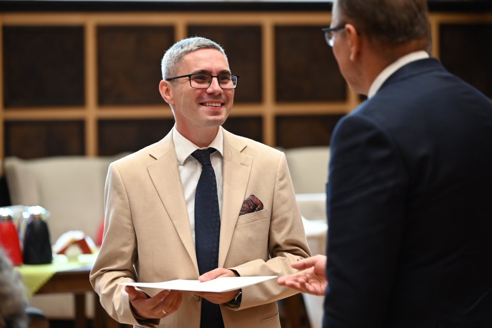 Na pierwszym planie znajdują się dwie osoby. Jeden z mężczyzn nosi jasny garnitur i ciemny krawat, a drugi ma na sobie ciemny garnitur. Mężczyźni zdają się być zaangażowani w rozmowę – mężczyzna w jasnym garniturze trzyma kartkę papieru. Atmosfera jest formalna, ale przyjazna. Wyraz twarzy mężczyzny w jasnym garniturze sugeruje zadowolenie lub satysfakcję. Tło jest wnętrzem budynku, z drewnianymi panelami i stołami. Widać również inne przedmioty, jak np. termosy. Na podstawie oświetlenia można przypuszczać, że zdjęcie zostało zrobione w ciągu dnia.