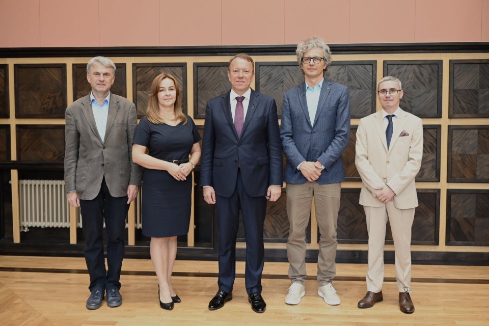 Na pierwszym planie znajduje się pięć osób, które stoją obok siebie. Wszyscy są elegancko ubrani, sugerując formalną okazję. Kobieta stoi pomiędzy czterema mężczyznami. W tle widoczna jest ściana wykończona drewnianymi panelami w ciemnych odcieniach. Na dole ściany widać również grzejnik. Osoby na zdjęciu stoją blisko siebie i patrzą w stronę obiektywu, co sugeruje, że pozują do fotografii grupowej. Atmosfera jest formalna, ale spokojna. Zdjęcie zostało zrobione w dobrze oświetlonym wnętrzu, co można wnioskować po jasnym oświetleniu sceny. Nie ma widocznych oznak sugerujących konkretną porę dnia czy roku.