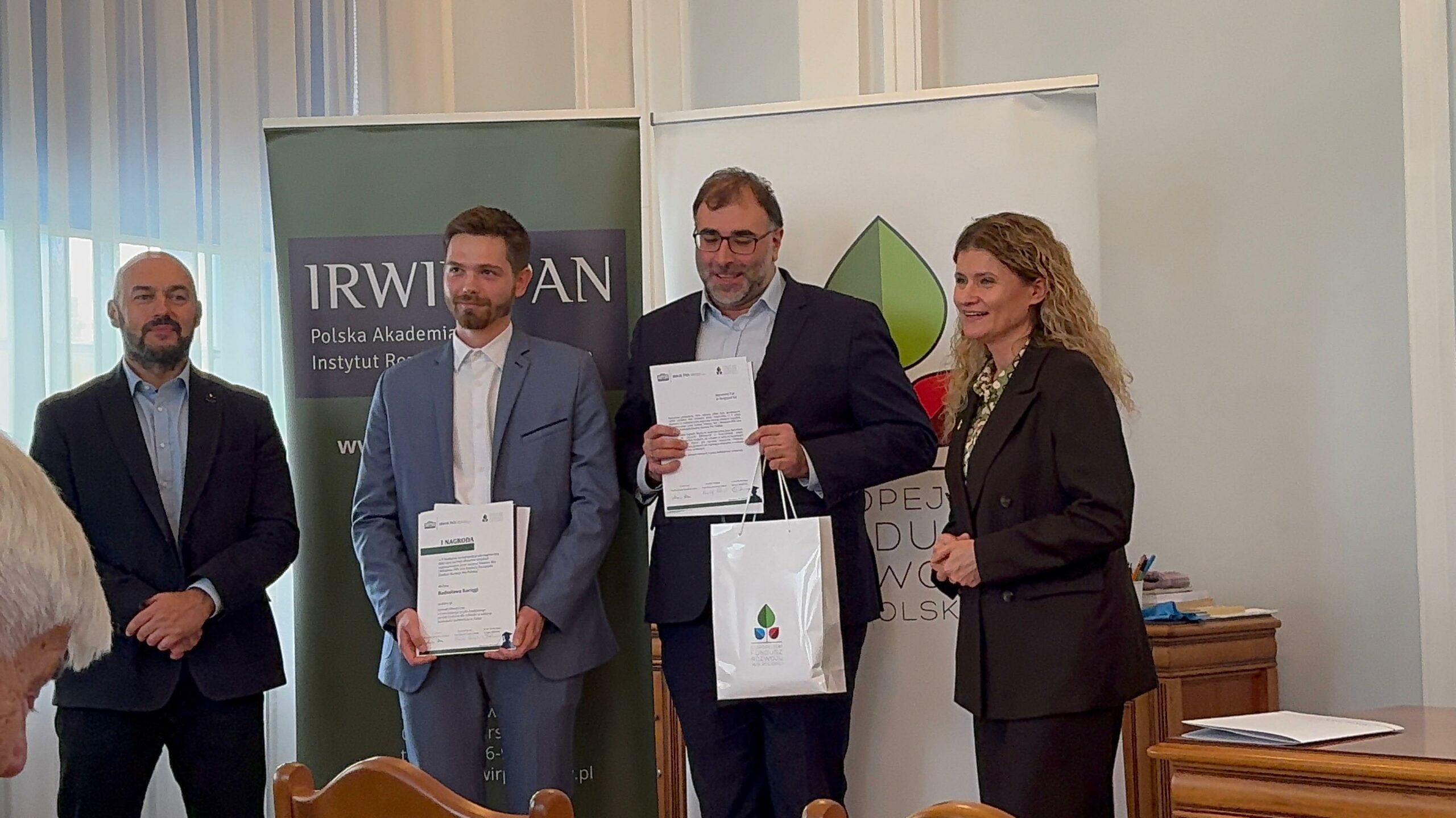Zdjęcie przedstawia formalne wydarzenie odbywające się w pomieszczeniu, prawdopodobnie ceremonię wręczenia nagród lub wyróżnień. W pierwszym planie stoją cztery osoby w eleganckich strojach — jedna z nich trzyma certyfikat i torbę z logo, a pozostali patrzą w jej stronę z uśmiechem. W tle widoczne są dwa banery z logo i nazwami „IRW” oraz „Polska Akademia Nauk”, co wskazuje na charakter naukowy lub instytucjonalny wydarzenia. Atmosfera jest uroczysta i profesjonalna, podkreślająca moment uznania lub osiągnięcia.