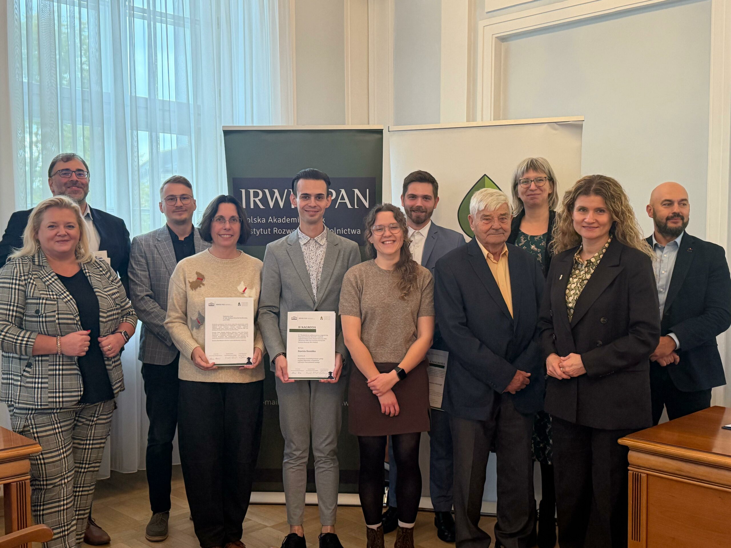 Na zdjęciu widać grupę dziesięciu osób pozujących do wspólnej fotografii w pomieszczeniu, prawdopodobnie podczas oficjalnego wydarzenia lub ceremonii wręczenia nagród. Uczestnicy — kobiety i mężczyźni w eleganckich strojach — stoją w dwóch rzędach i patrzą w obiektyw, uśmiechając się. W centrum znajdują się dwie osoby trzymające certyfikaty, co sugeruje wyróżnienie lub zakończenie projektu. W tle widoczny jest baner z napisem „IRW PAN” i „Polska Akademia Nauk”, wskazujący na związek wydarzenia z instytucją naukową. Atmosfera zdjęcia jest uroczysta, ale jednocześnie przyjazna i pełna dumy.