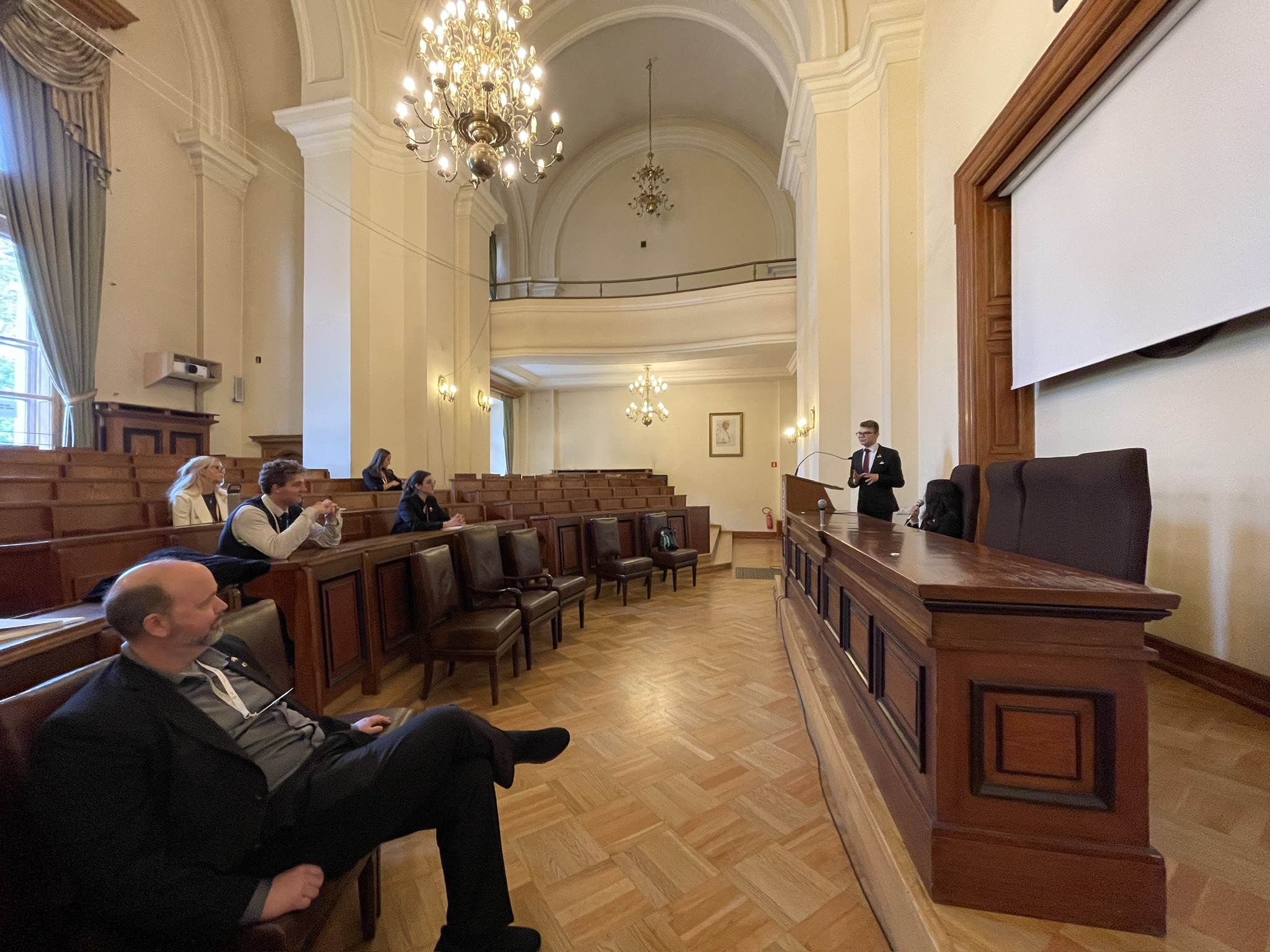 Na zdjęciu znajduje się przestronna, elegancka aula z wysokim sklepieniem i dużymi żyrandolami. Po prawej stronie, przy drewnianym stole prezydialnym, stoi mężczyzna przemawiający do mikrofonu ustawionego na mównicy. W ławach po lewej stronie sali siedzi kilka osób, słuchających wystąpienia. Wnętrze jest jasne, z dużymi oknami i ciężkimi zasłonami, a na podłodze ułożony jest parkiet. Całość utrzymana jest w klasycznym, uniwersyteckim stylu, podkreślając oficjalny charakter wydarzenia.