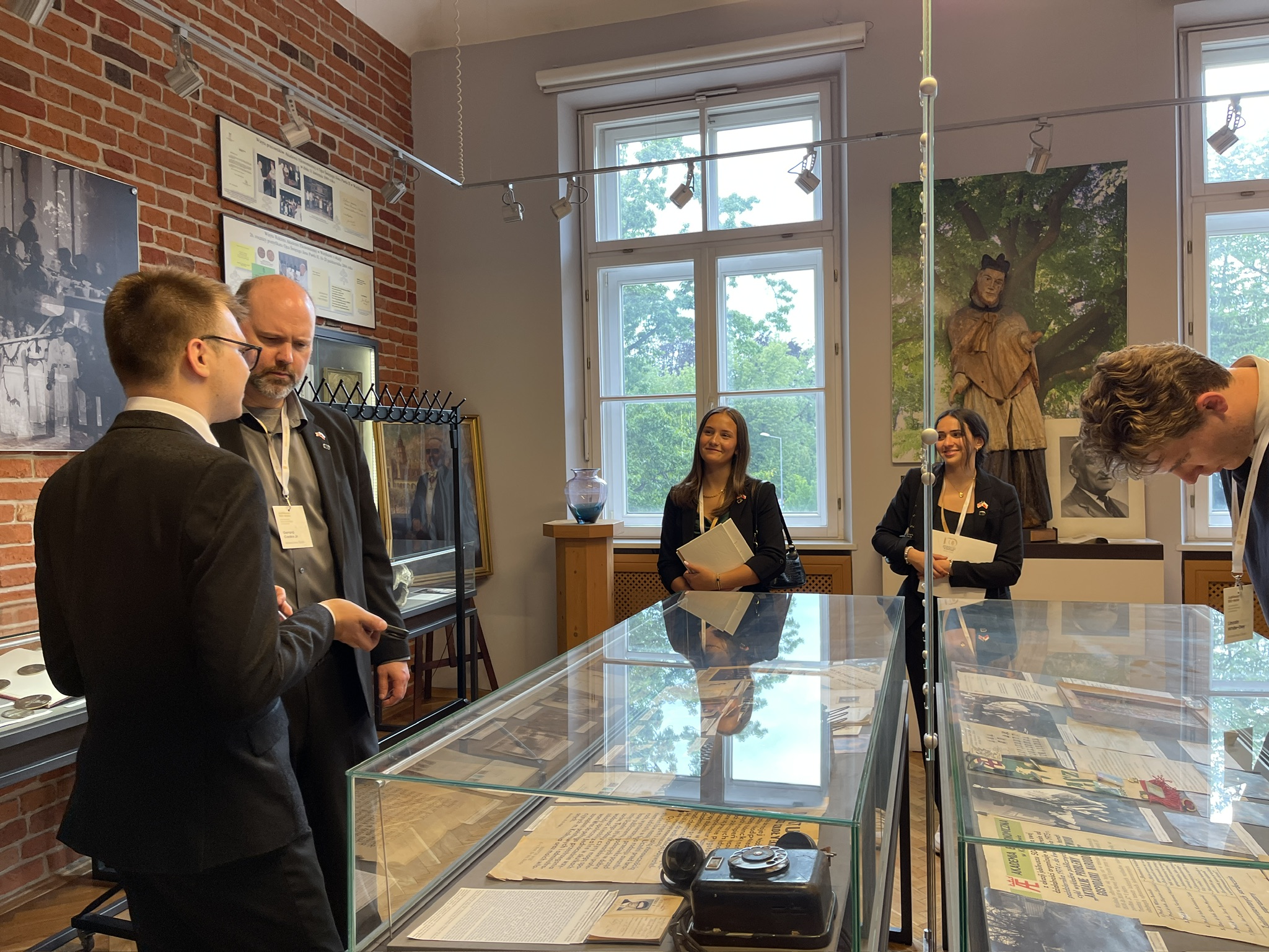 Na zdjęciu znajduje się pięć osób w muzealnym wnętrzu z ceglanymi ścianami. Dwie osoby stoją po lewej stronie, zwrócone do siebie w rozmowie, natomiast trzy pozostałe osoby znajdują się po prawej stronie, patrząc na eksponaty lub trzymając w rękach broszury. W pomieszczeniu dominują duże okna, przez które widać zieleń na zewnątrz. W centralnej części zdjęcia widoczne są przeszklone gabloty z dokumentami i starym aparatem fotograficznym. Na ścianach wiszą fotografie i obrazy, a w głębi sali stoi rzeźba kobiety w historycznym stroju oraz popiersie. Całość wnętrza utrzymana jest w klimacie historycznym i muzealnym.