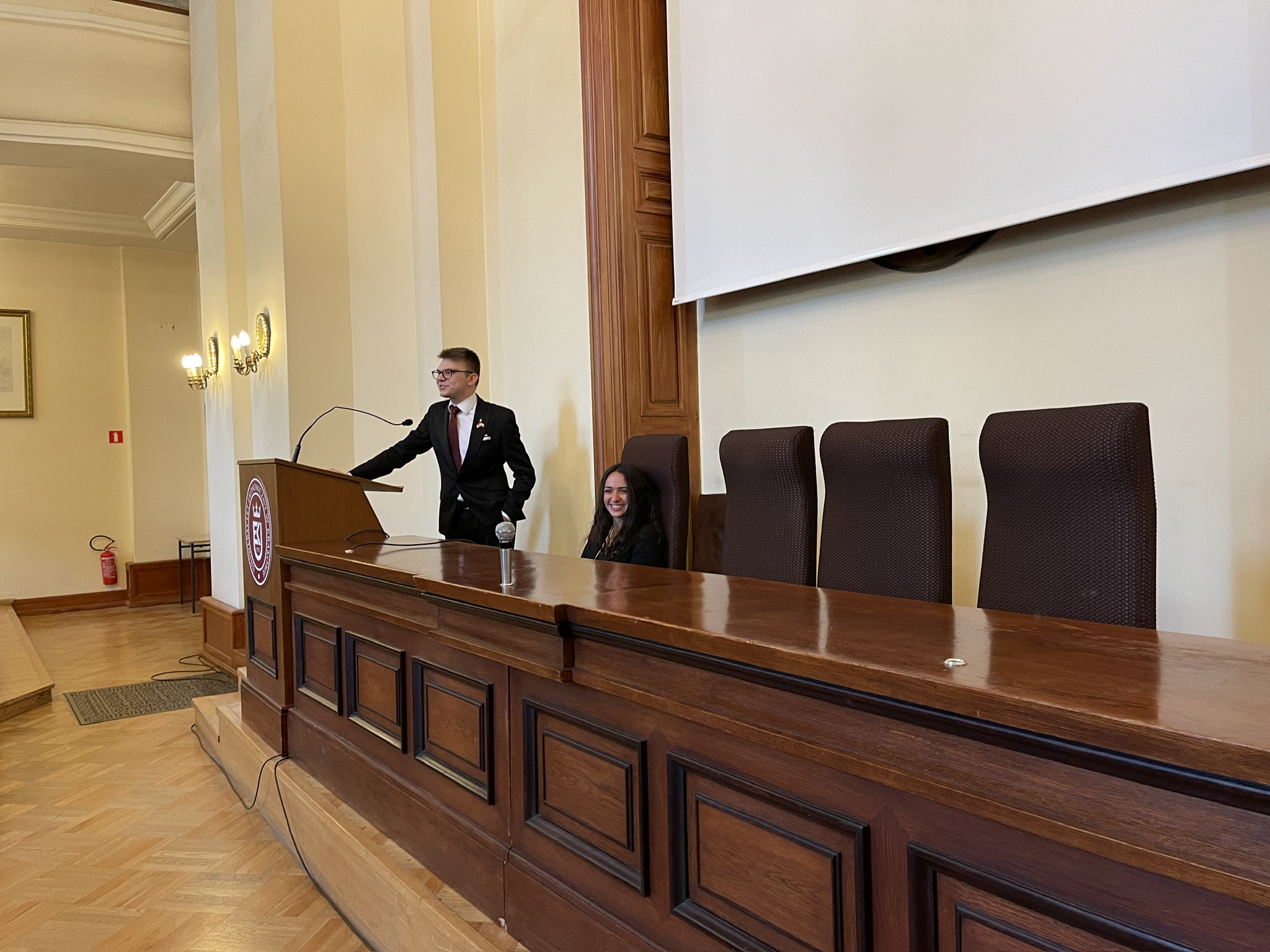 Na zdjęciu widoczna jest Stara Aula  z drewnianym stołem prezydialnym ustawionym na podwyższeniu. Przy stole stoi mężczyzna w garniturze, przemawiający do mikrofonu ustawionego na mównicy z logo uczelni. Obok niego, przy stole, siedzi druga osoba. W tle znajduje się duży, opuszczony ekran projekcyjny oraz cztery wysokie krzesła ustawione przy stole. Ściany sali są jasne, ozdobione kinkietami i drewnianymi panelami. Na podłodze ułożony jest parkiet, a w lewym rogu pomieszczenia widoczna jest gaśnica oraz fragment ramki z obrazem. Całość wnętrza utrzymana jest w eleganckim, klasycznym stylu.