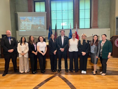 Na zdjęciu znajduje się grupa jedenastu osób ustawionych w jednym rzędzie w przestronnej sali konferencyjnej. W tle widoczne są duże, prostokątne okna oraz ekran projekcyjny, na którym wyświetlona jest prezentacja z napisem „BEYOND THE DECADES: CELEBRATING A JOINT HISTORY, INSPIRING THE FUTURE”. Za grupą stoją flagi, w tym flaga Unii Europejskiej oraz Polski. Po prawej stronie widoczna jest mównica z logo uczelni. Podłoga wykonana jest z jasnego drewna, a całość wnętrza utrzymana jest w eleganckim, oficjalnym stylu.