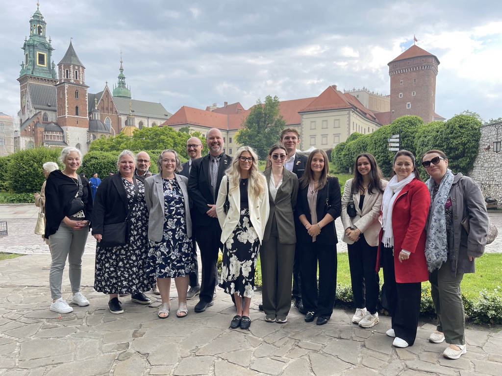 Na zdjęciu znajduje się grupa jedenastu osób stojących na dziedzińcu Zamku Królewskiego na Wawelu w Krakowie. W tle widoczne są charakterystyczne zabudowania zamku, w tym wieże katedry wawelskiej oraz ceglany budynek z okrągłą basztą. Osoby ustawione są w jednym rzędzie na kamiennej nawierzchni, otoczone zielenią i historyczną architekturą. Pogoda jest pochmurna, ale jasna, co podkreśla wyjątkowy klimat miejsca. Całość zdjęcia utrzymana jest w eleganckim, pamiątkowym charakterze.