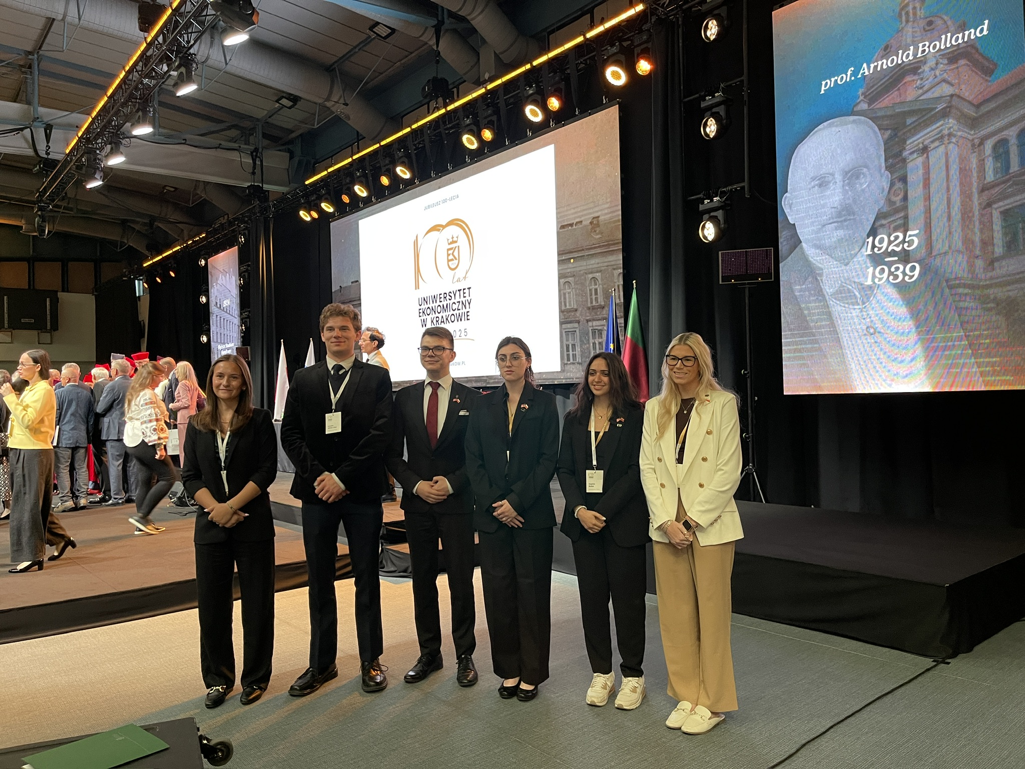 Na zdjęciu znajduje się sześć osób stojących w rzędzie na tle sceny podczas uroczystości na Uniwersytecie Ekonomicznym w Krakowie. W tle widoczny jest duży ekran z jubileuszowym logo uczelni oraz fragmentem prezentacji poświęconej prof. Arnoldowi Bollandowi. Po prawej stronie ekranu stoją flagi, a całość oświetlona jest profesjonalnym światłem scenicznym. Po lewej stronie, na scenie, widać grupę osób zgromadzonych w trakcie wydarzenia. Wnętrze sali jest przestronne i nowoczesne, a atmosfera zdjęcia jest oficjalna i uroczysta