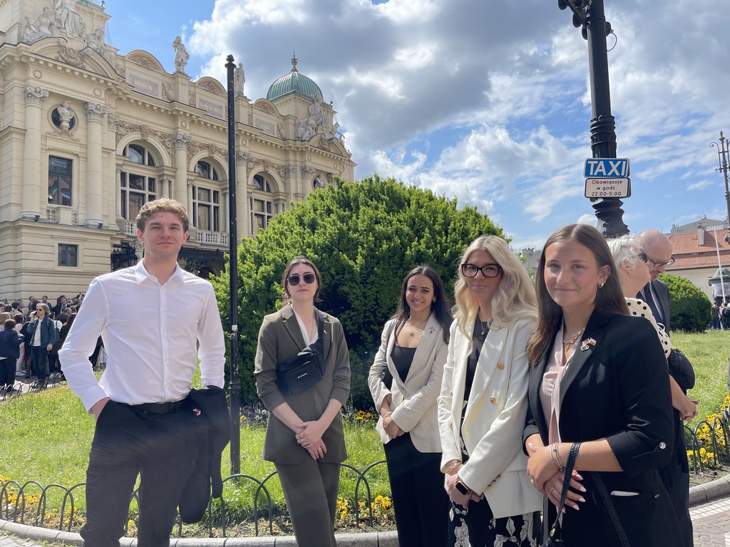 Na zdjęciu znajduje się sześć osób stojących na tle zabytkowego budynku Teatru im. Juliusza Słowackiego w Krakowie. Osoby ustawione są w dwóch rzędach na zielonym skwerze, otoczonym niskim, metalowym ogrodzeniem i kwiatami. W tle widoczna jest fasada teatru z charakterystyczną kopułą oraz liczna grupa ludzi zgromadzonych przed wejściem.  Nad całością rozpościera się niebo z częściowo zachmurzonym, słonecznym niebem, co nadaje zdjęciu jasny i pogodny charakter.