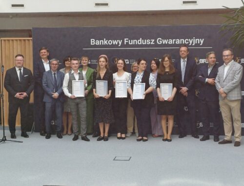 Grupowe zdjęcie uczestników i organizatorów wydarzenia z okazji wręczenia nagród lub certyfikatów przez Bankowy Fundusz Gwarancyjny. W pierwszym rzędzie stoi sześć osób trzymających dyplomy, za nimi grupa przedstawicieli w formalnych strojach. W tle ścianka z logo Bankowego Funduszu Gwarancyjnego.