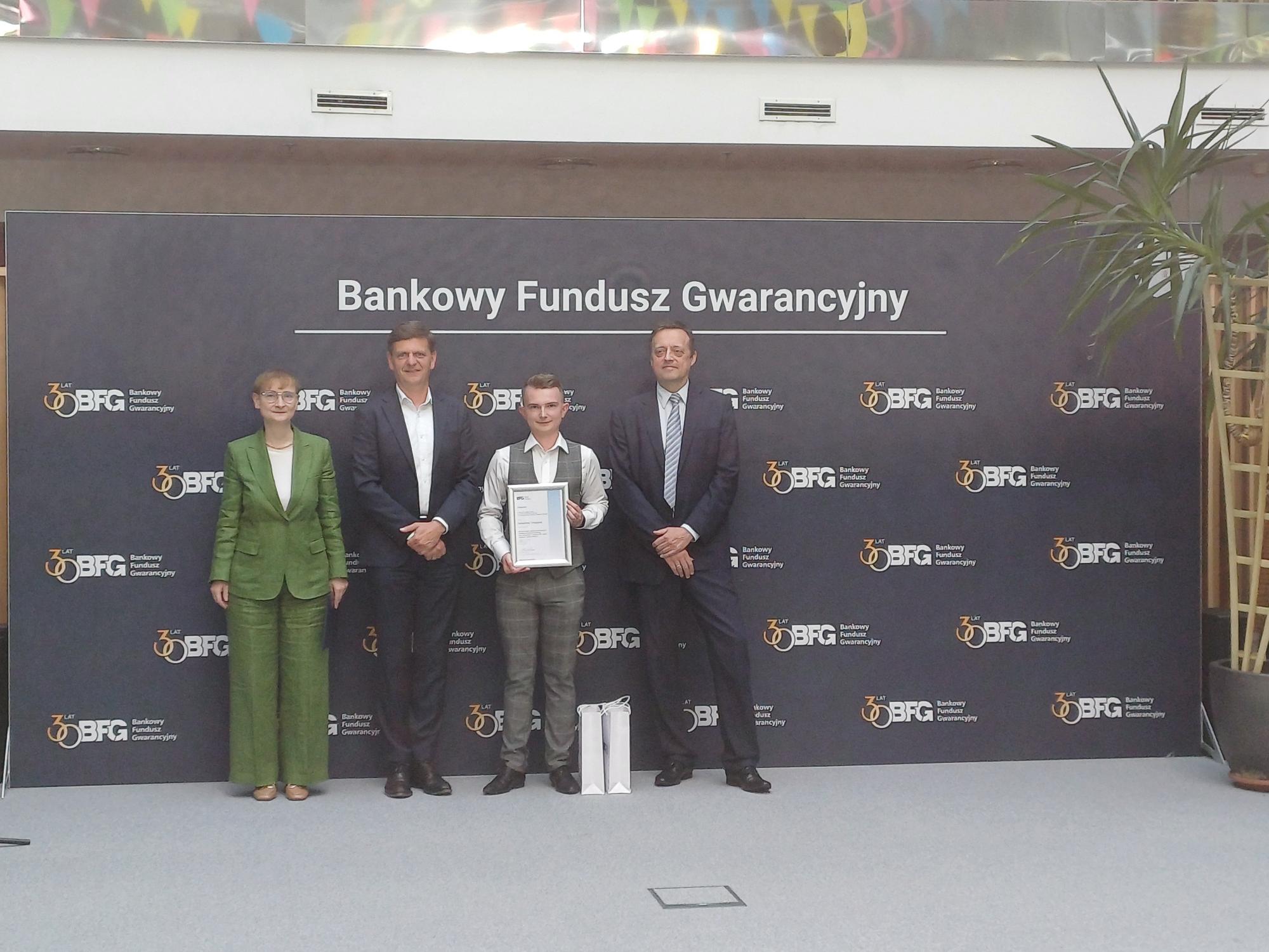 Indywidualne zdjęcie laureata trzymającego dyplom, stojącego na tle ścianki z logo Bankowego Funduszu Gwarancyjnego. Po jego bokach stoją trzej mężczyźni i jedna kobieta w eleganckich strojach. Na podłodze przed laureatem znajdują się dwie białe torby z upominkami.