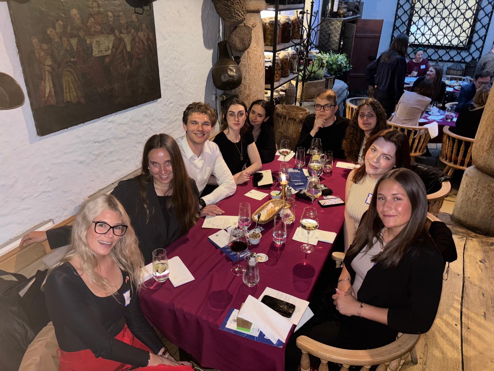 Na zdjęciu widać grupę dziewięciu osób siedzących przy prostokątnym stole nakrytym bordowym obrusem w restauracyjnym wnętrzu o historycznym charakterze. Na stole stoją kieliszki z wodą, talerzyki, karafka oraz koszyk z pieczywem. W tle widoczne są inne stoliki z gośćmi, elementy drewnianego wystroju oraz półki z dekoracjami. Na ścianie wisi duży obraz przedstawiający scenę biesiadną. Uczestnicy spotkania są elegancko ubrani, a atmosfera wydaje się formalna, sugerująca kolację integracyjną lub wydarzenie towarzyskie o oficjalnym charakterze.