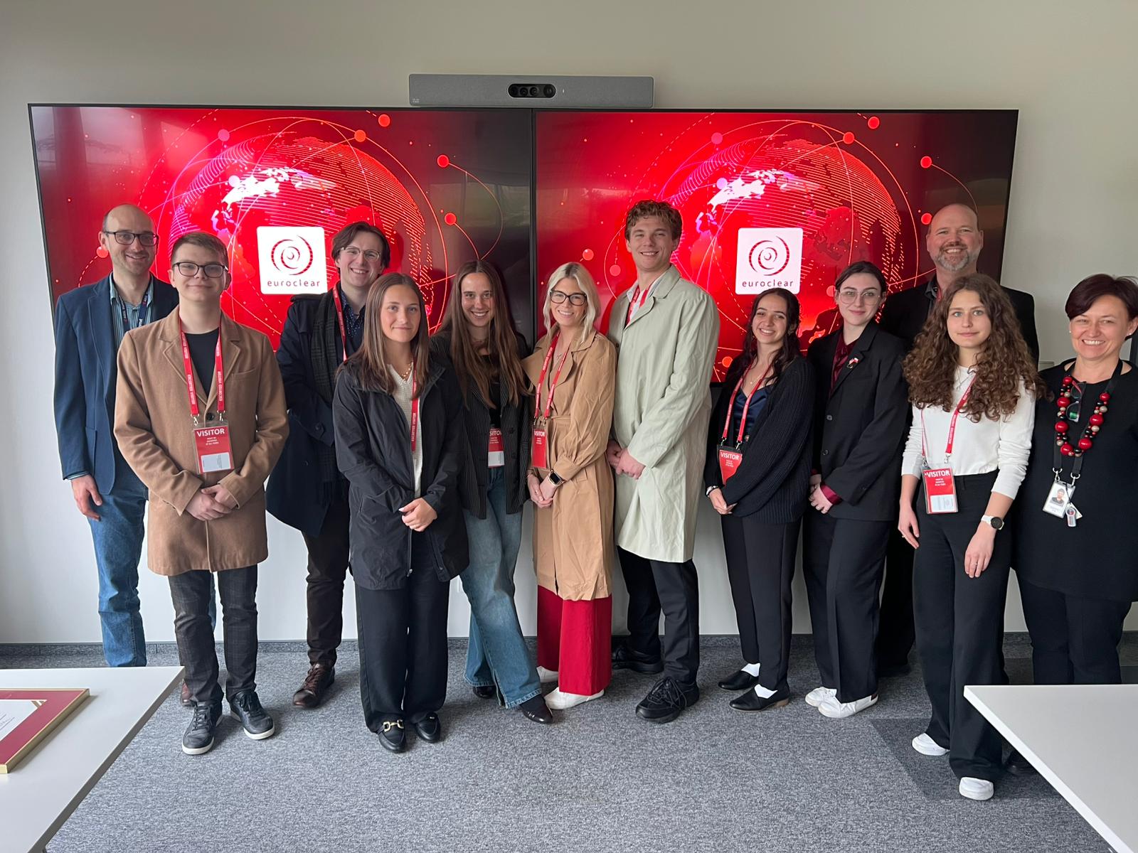 Na zdjęciu widoczna jest grupa jedenastu osób stojących w nowoczesnym pomieszczeniu biurowym. W tle znajdują się dwa duże ekrany z czerwonym graficznym motywem przedstawiającym mapę świata oraz logo firmy Euroclear. Wszyscy uczestnicy mają na szyjach czerwone smycze z identyfikatorami typu “VISITOR”, co sugeruje, że są gośćmi na firmowym wydarzeniu lub wycieczce. Ubrania osób są półformalne – dominują płaszcze, marynarki, eleganckie spodnie oraz buty. Atmosfera zdjęcia jest oficjalna i profesjonalna, a tło oraz identyfikatory wskazują na wizytę w międzynarodowej instytucji.