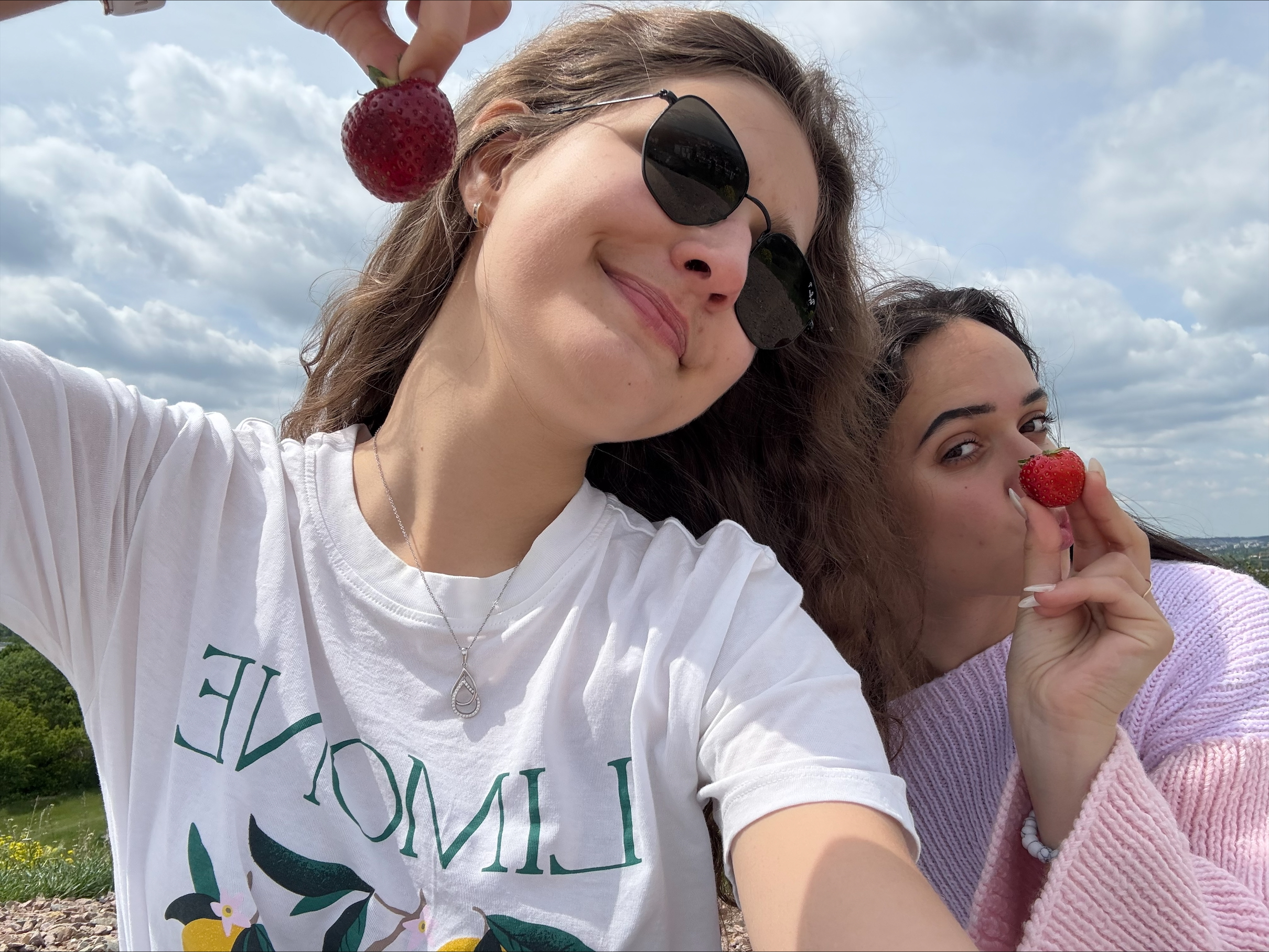 Na zdjęciu dwie osoby pozują na tle krajobrazu z zielonymi polami i niebem pokrytym chmurami. Jedna z nich trzyma w górze dużą czerwoną truskawkę, druga również trzyma truskawkę przy twarzy. Osoba po lewej stronie ma na sobie białą koszulkę z nadrukiem cytryn i napisem „LIMONE” oraz srebrny naszyjnik, a osoba po prawej ubrana jest w jasnoróżowy sweter i ma na nadgarstku białą bransoletkę. Zdjęcie zostało wykonane na zewnątrz, w pogodny dzień, co podkreślają jasne kolory i naturalne światło. Całość sprawia wrażenie spontanicznej, letniej chwili podczas wspólnego spaceru.