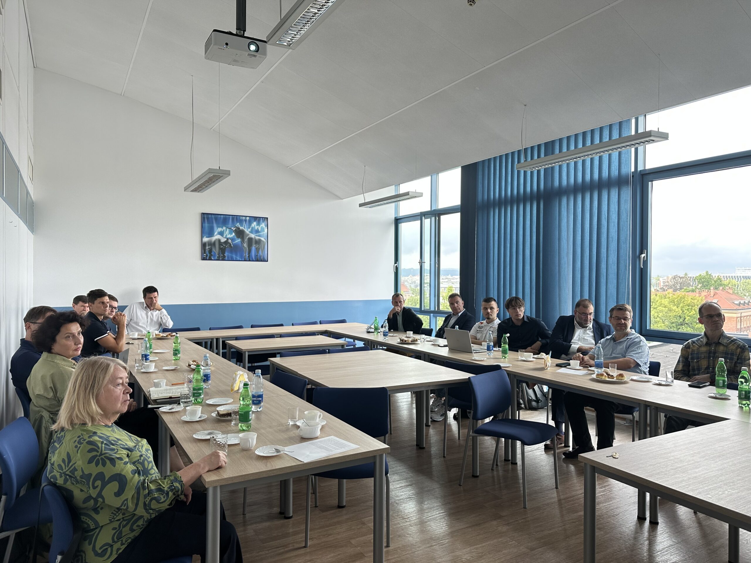 Na pierwszym planie widzimy grupę osób siedzących przy stołach w kształcie litery U. Stoły są ustawione w jasnym pomieszczeniu. Na stołach znajdują się napoje, filiżanki i talerze z przekąskami. Ludzie wyglądają na skupionych. W tle dostrzec można duże okna z niebieskimi zasłonami, przez które widać drzewa i fragment miejskiego krajobrazu, co sugeruje, że pokój znajduje się na wyższych piętrach budynku. Na ścianie widnieje obraz lub plakat przedstawiający fragment natury. Atmosfera jest formalna i profesjonalna. Część osób wygląda na zaangażowanych w rozmowę lub słuchanie prezentacji. Widać również projektor podwieszony na suficie. Pora dnia sugerowana przez oświetlenie i widoczność za oknami to późne rano lub wczesne popołudnie. Pomieszczenie jest dobrze oświetlone naturalnym światłem.