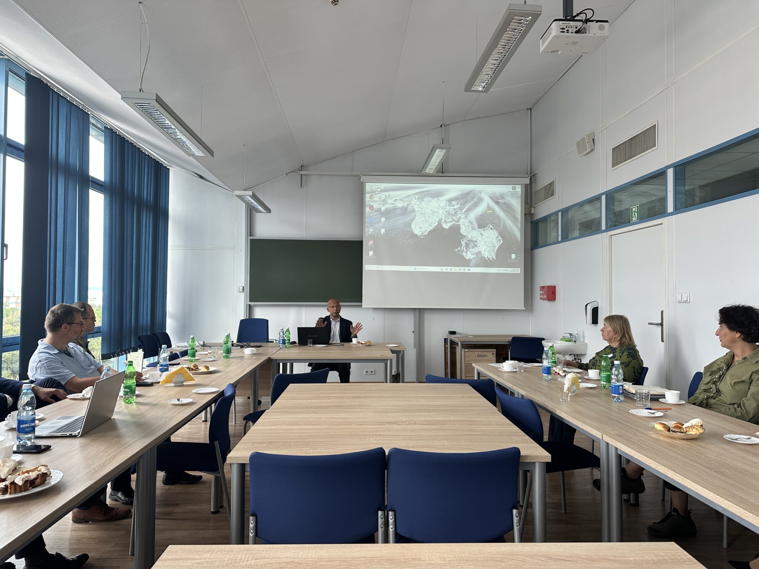 Na pierwszym planie widzimy salę konferencyjną z ustawionymi w kształt litery "U" drewnianymi stołami. Na stołach znajdują się butelki z wodą, filiżanki do kawy, talerze z przekąskami oraz laptopy. Po bokach siedzą ludzie. W tle, na ścianie, widzimy ekran projekcyjny z wyświetlonym obrazem mapy satelitarnej. Przed ekranem stoi mężczyzna przy laptopie. Na ścianie jest także widoczna zielona tablica oraz projektor zawieszony pod sufitem. Osoby na zdjęciu siedzą i patrzą w kierunku prezentera. Pomieszczenie wydaje się być dobrze oświetlone, co sugeruje, że zdjęcie wykonano w ciągu dnia. Przez okno po lewej stronie wpada naturalne światło. Można zauważyć, że jest to wnętrze nowoczesnego budynku.