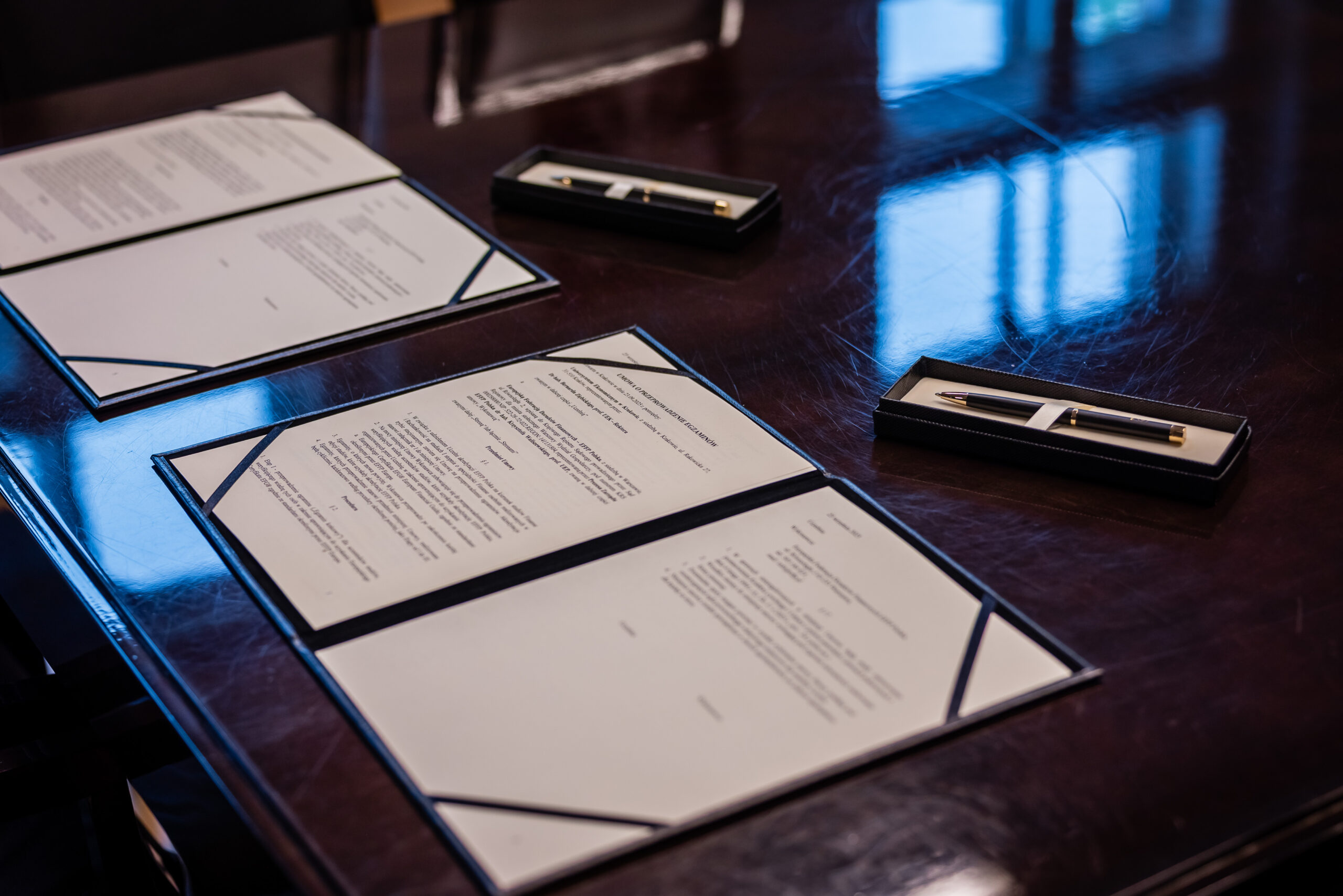 Na pierwszym planie zdjęcia widzimy stół, na którym leżą dokumenty oraz pióra w eleganckich etui. Dokumenty są ułożone w otwarty sposób, co sugeruje gotowość do podpisania lub przeglądania. Każdy dokument znajduje się w ozdobnym folderze, co dodaje formalnego charakteru oświetleniu. W tle widoczne jest odbicie światła, które może pochodzić od okien, sugerując, że zdjęcie zostało zrobione w ciągu dnia, prawdopodobnie rano lub wczesnym popołudniem. Atmosfera jest formalna i profesjonalna, co może wskazywać na miejsce takie jak sala konferencyjna lub biuro. Obiekty są w interakcji pośredniej - przygotowane do podpisania dokumenty dają wrażenie oczekiwania na ważne spotkanie lub uroczystość biznesową. Pióra leżące obok dokumentów sugerują, że podpisanie może być wkrótce. Zdjęcie jest realistyczne; wykonane z dużą dbałością o detale, co podkreśla elegancję i powagę sytuacji. Brak jest bezpośrednich ludzkich postaci, co kieruje uwagę tylko na przedmioty. Całość oddaje atmosferę koncentracji i profesjonalizmu.