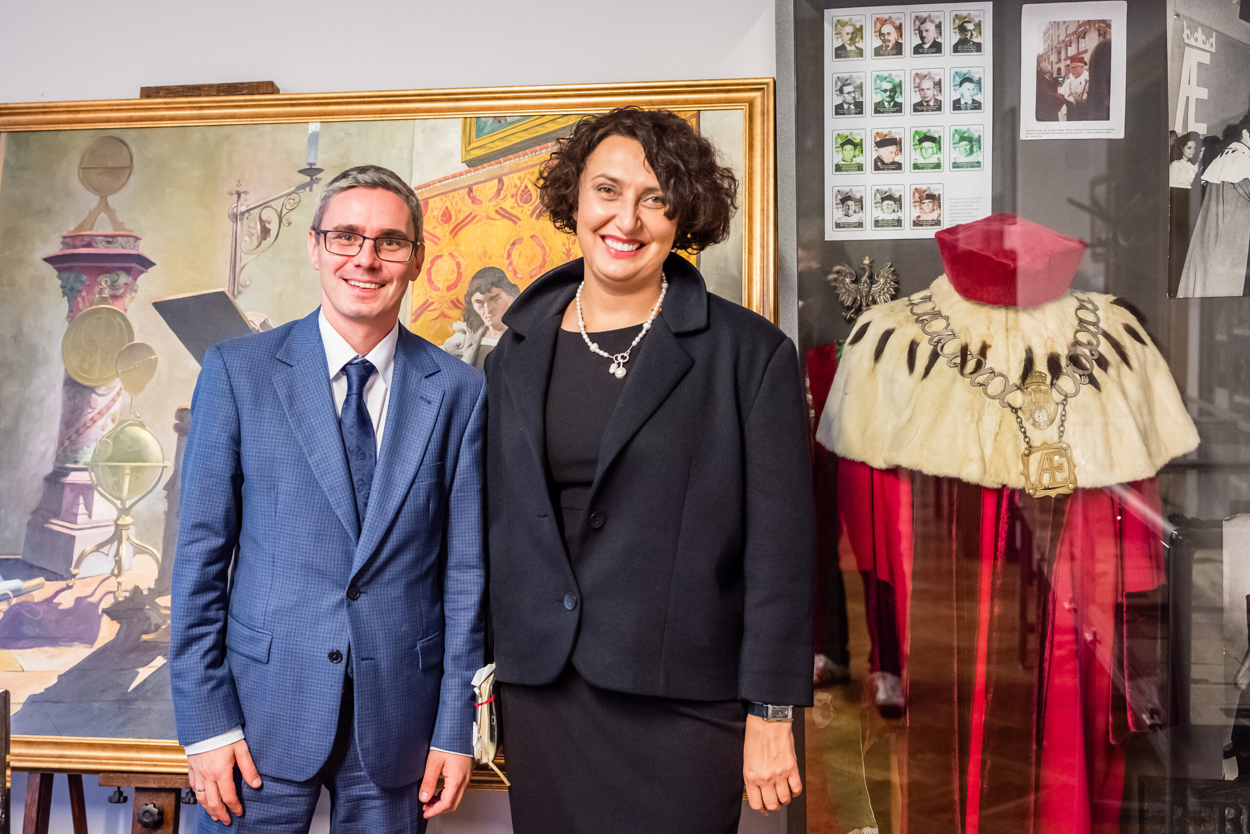 Na pierwszym planie widoczne są dwie osoby ubrane formalnie. Mężczyzna po lewej stronie ma na sobie niebieski garnitur, białą koszulę i granatowy krawat. Kobieta po prawej nosi czarny strój składający się z marynarki i sukienki. Oboje uśmiechają się i stoją blisko siebie. W tle, na ścianie, widoczny jest duży obraz w złotej ramie, przedstawiający klasyczny, ozdobny motyw z elementami architektury i symboliki. Obok znajduje się gablotka z ceremonialnym strojem w czerwono-białych kolorach, ozdobionym srebrnym łańcuchem, co może sugerować akademickie lub urzędowe znaczenie. Atmosfera zdjęcia jest formalna, sugerująca akademickie lub zawodowe wydarzenie. Ciepłe kolory obrazu i ceremonialnego stroju nadają scenie elegancki charakter. Wystrój wnętrza i sposób ubrania osób mogą sugerować, że zdjęcie zostało wykonane wewnątrz budynku, prawdopodobnie w instytucji edukacyjnej lub kulturalnej. Na podstawie stroju nie można dokładnie określić pory dnia czy roku. Zdjęcie jest realistyczne, prawdopodobnie wykonane aparatem fotograficznym.
