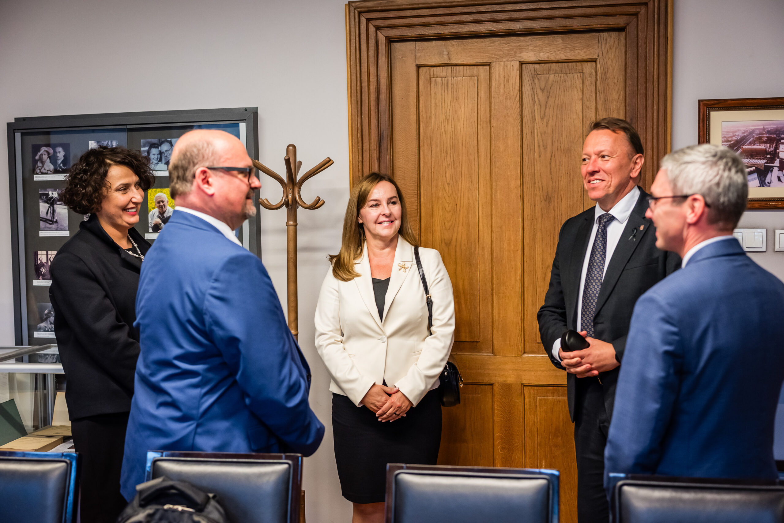 Na pierwszym planie zdjęcia widoczne jest pięć osób stojących w niewielkiej grupie, najwyraźniej rozmawiających ze sobą. Wszyscy są elegancko ubrani – w garnitury i eleganckie stroje, sugerujące formalne spotkanie lub wydarzenie biznesowe. W tle znajduje się jasne, drewniane drzwi, a obok nich wieszak na ubrania. Na ścianie można zauważyć ramki z fotografiami oraz częściowo widoczną szafkę lub gablotę. Atmosfera w pomieszczeniu jest prawdopodobnie formalna, ale również serdeczna, o czym świadczą uśmiechy osób na zdjęciu. Osoby na zdjęciu wydają się być zaangażowane w rozmowę. Emocje wydają się być pozytywne — uśmiechy i postawa ciała sugerują przyjazny ton rozmowy. Pomieszczenie wygląda na biurowe lub konferencyjne. Nie widać bezpośrednio żadnych oznak wskazujących na porę dnia lub roku.
