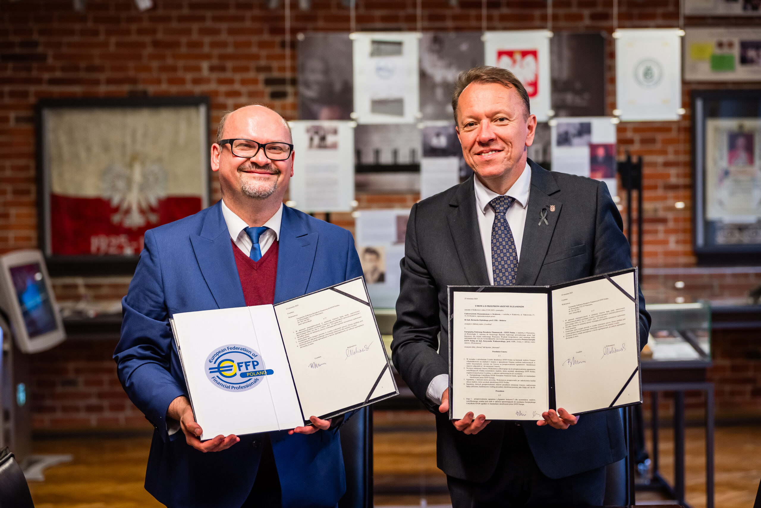 Na pierwszym planie widzimy dwóch mężczyzn ubranych elegancko w garnitury. Każdy z nich trzyma otwarty dokument. Mężczyźni wyglądają na zadowolonych, a ich postawa wyraża profesjonalizm i sukces. W tle znajduje się ściana z cegieł z licznymi zawieszonymi zdjęciami oraz dokumentami. Jest to przestrzeń formalna, zorganizowana i dobrze oświetlona, co dodaje atmosferze powagi i oficjalności. Na fotografii nie widzimy bezpośrednich wskazówek dotyczących pory dnia, ale oświetlenie jest jasne i wydaje się, że zdjęcie zostało zrobione w ciągu dnia.