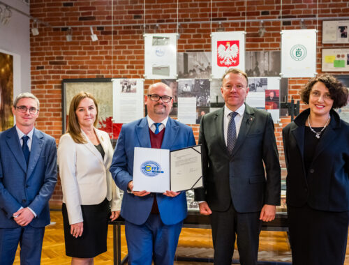 W przedstawionym obrazie możemy zobaczyć grupę pięciu osób stojących w pomieszczeniu o wyglądzie muzealnym. W centralnej części obrazu znajduje się mężczyzna trzymający w rękach dyplom. Wokół niego znajdują się cztery inne osoby - dwie z lewej i dwie z prawej strony. Tło stanowią ceglaste ściany z zawieszonymi dokumentami i fotografiami, co sugeruje, że miejsce to ma charakter historyczny lub edukacyjny. W tle widoczne są symbole takie jak godło Polski, co może wskazywać na oficjalny charakter instytucji. Osoby na zdjęciu są ubrane w eleganckie stroje. Wszyscy patrzą w stronę aparatu, a ich wyraz twarzy jest profesjonalny, co wskazuje na oficjalne spotkanie bądź ceremonię wręczenia nagród. Atmosfera obrazu jest formalna i profesjonalna. Wystrój pomieszczenia oraz obecność dokumentów wskazują na instytucjonalność miejsca. Czas dnia nie jest ściśle określony, ale jasne oświetlenie sugeruje, że zdjęcie mogło być wykonane w ciągu dnia.