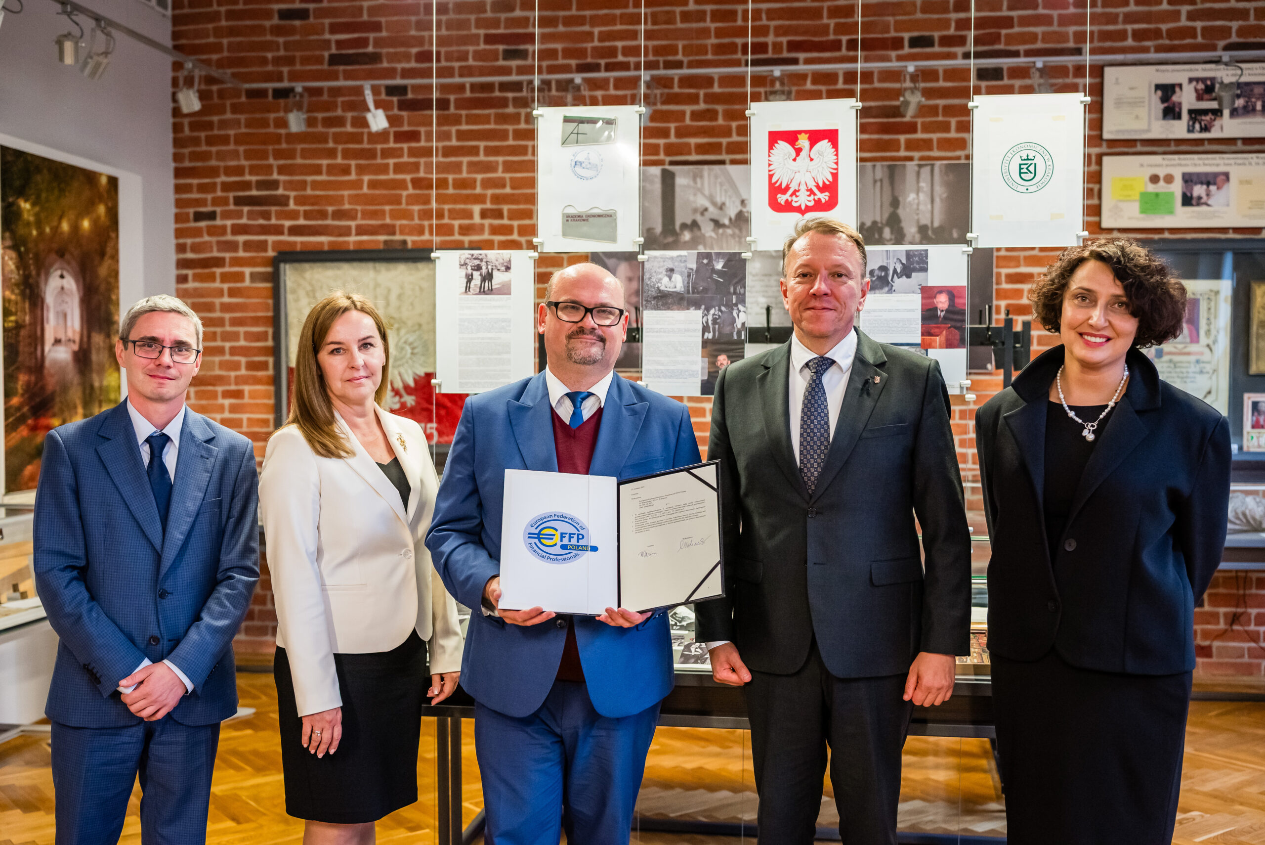 W przedstawionym obrazie możemy zobaczyć grupę pięciu osób stojących w pomieszczeniu o wyglądzie muzealnym. W centralnej części obrazu znajduje się mężczyzna trzymający w rękach dyplom. Wokół niego znajdują się cztery inne osoby - dwie z lewej i dwie z prawej strony. Tło stanowią ceglaste ściany z zawieszonymi dokumentami i fotografiami, co sugeruje, że miejsce to ma charakter historyczny lub edukacyjny. W tle widoczne są symbole takie jak godło Polski, co może wskazywać na oficjalny charakter instytucji. Osoby na zdjęciu są ubrane w eleganckie stroje. Wszyscy patrzą w stronę aparatu, a ich wyraz twarzy jest profesjonalny, co wskazuje na oficjalne spotkanie bądź ceremonię wręczenia nagród. Atmosfera obrazu jest formalna i profesjonalna. Wystrój pomieszczenia oraz obecność dokumentów wskazują na instytucjonalność miejsca. Czas dnia nie jest ściśle określony, ale jasne oświetlenie sugeruje, że zdjęcie mogło być wykonane w ciągu dnia.