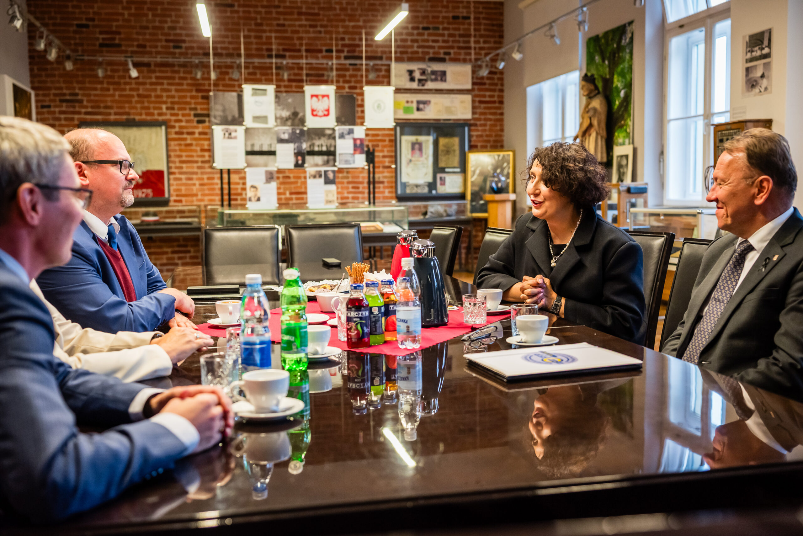 W przedstawionym obrazie znajduje się scena spotkania. W centrum uwagi, na pierwszym planie, jest stół z wyłożonymi różnorodnymi napojami i filiżankami kawy. Na stole znajduje się również talerz z przekąskami oraz dokumenty. Wokół stołu siedzi pięć osób, które uczestniczą w oficjalnym spotkaniu. Nosi to znamiona formalnego spotkania, co sugerują eleganckie ubrania uczestników – garnitury i marynarki. Osoby te są zwrócone twarzami ku sobie, co może wskazywać na zaangażowanie w rozmowę. Atmosfera spotkania wydaje się być formalna, ale jednocześnie przyjazna i otwarta. W tle widzimy ceglaną ścianę z zawieszonymi na niej dokumentami oraz znakami. W pomieszczeniu znajduje się także duży okno, które wpuszcza naturalne światło, co sugeruje, że zdjęcie zostało zrobione w ciągu dnia. W tle widoczne są także inne elementy dekoracyjne, takie jak obrazy czy rzeźby, co może świadczyć o tym, że jest to przestrzeń użytku publicznego, może galeria lub miejsce o historycznym znaczeniu.