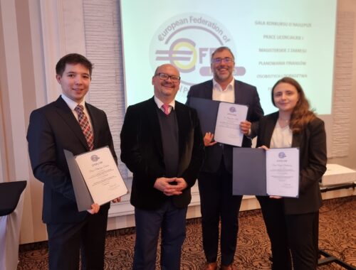 Na pierwszym planie zdjęcia widocznych jest czterech osób, trzy z nich trzymają dyplomy. Osoby te są elegancko ubrane. W tle widać ekran z logo i napisami. Sceneria wskazuje, że jest to wnętrze sali konferencyjnej. Atmosfera zdjęcia jest uroczysta i oficjalna, co podkreślone jest formalnym strojem obecnych osób i dyplomami, które trzymają. Stylizacje wnętrza oraz sposób ubioru wskazują na formalne wydarzenie. Nie ma widocznych oznak, które mogłyby dokładnie określić porę dnia czy roku, ale obecność światła sugeruje, że zdjęcie zostało zrobione w ciągu dnia.