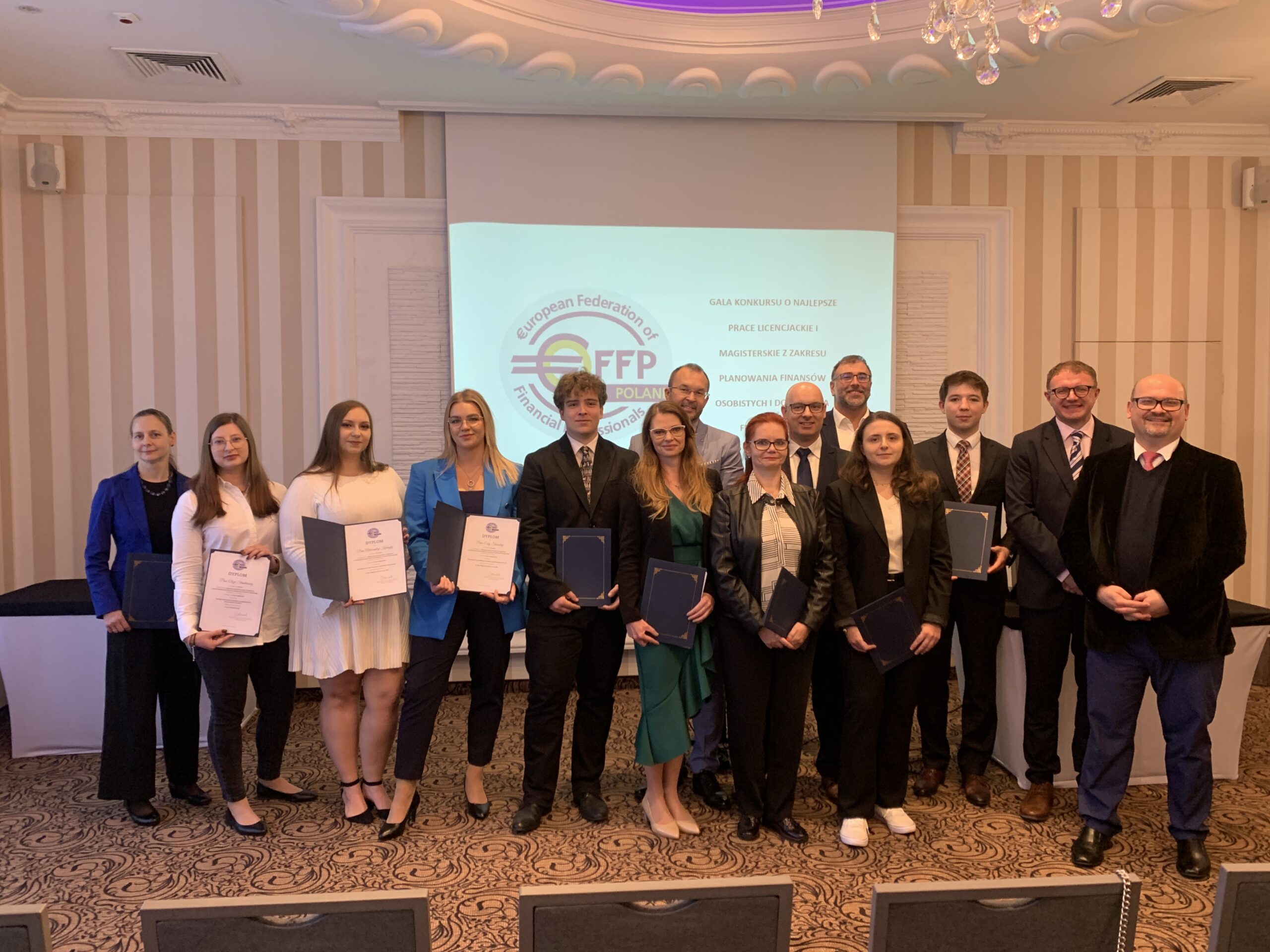 W przedstawionym obrazie widzimy grupę osób stojących w rzędzie na tle ekranu projekcyjnego, na którym wyświetlana jest prezentacja z logotypem organizacji. **Pierwszy plan:** - W centrum zdjęcia znajduje się kilkanaście osób ubranych w formalne stroje, takie jak marynarki, koszule i eleganckie sukienki. Niektórzy trzymają dokumenty lub teczki w dłoniach. **Tło:** - W tle widoczny jest ekran z logo oraz tekstem, jasne oświetlenie oraz eleganckie, klasyczne wnętrze. **Główne obiekty/osoby:** - Grupa ubranych formalnie osób; niektórzy z nich uśmiechają się. **Działania/interakcja:** - Osoby stojące w rzędzie blisko siebie, co wskazuje na wspólne uczestnictwo w oficjalnej ceremonii lub gali. **Emocje/atmosfera:** - Atmosfera jest uroczysta i oficjalna, z nutą radości i dumy, co podkreślają uśmiechy uczestników. **Lokalizacja/ustawienie:** - Sala konferencyjna. **Czas dnia/pora roku:** - Nie można jednoznacznie określić pory dnia ani roku na podstawie samej fotografii.