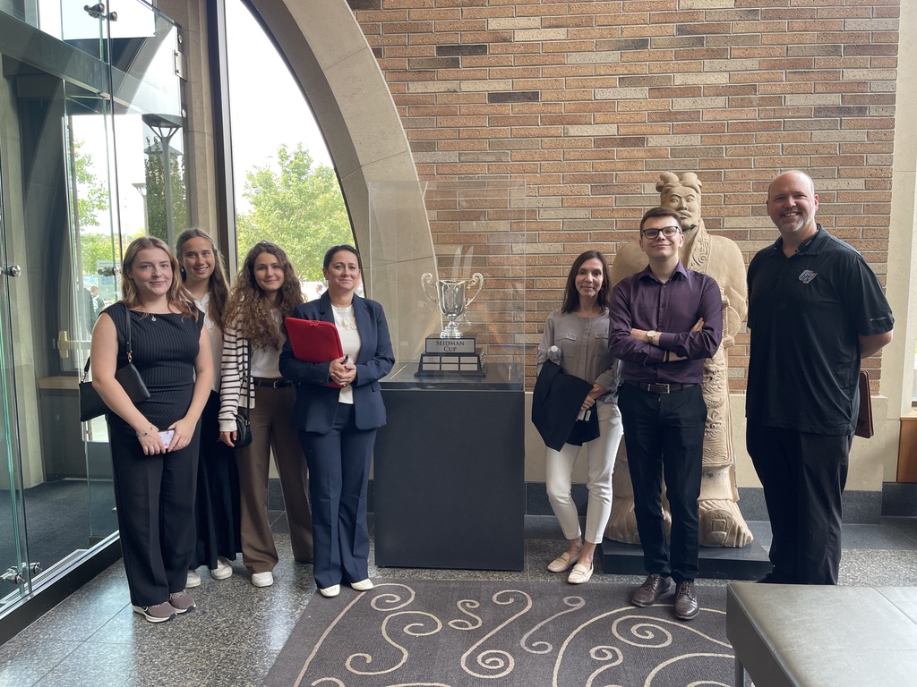 Na zdjęciu znajduje się grupa siedmiu osób stojących w eleganckim holu Seidman College of Business, GVSU. W centralnej części, na czarnym postumencie, widoczny jest srebrny puchar w szklanej gablocie. Za grupą znajduje się ściana z jasnobrązowej cegły oraz kamienna rzeźba postaci w tradycyjnym stroju. Po lewej stronie widać duże przeszklone drzwi, przez które wpada naturalne światło. Podłoga pokryta jest ciemnym kamieniem z dekoracyjnym, jasnym wzorem.