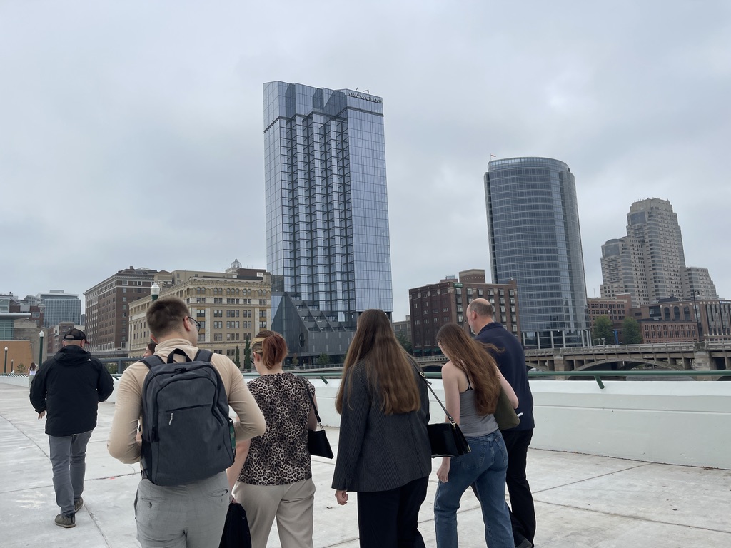 Na zdjęciu widać grupę sześciu osób idących szerokim chodnikiem przez most w centrum miasta. Osoby są ubrane swobodnie, a niektóre z nich mają na plecach plecaki lub torby. W tle dominują nowoczesne, wysokie budynki o szklanych elewacjach oraz starsze, ceglane kamienice. Niebo jest zachmurzone, a otoczenie miejskie sprawia wrażenie przestronnego i nowoczesnego. Po prawej stronie kadru widoczna jest rzeka oraz balustrada mostu.