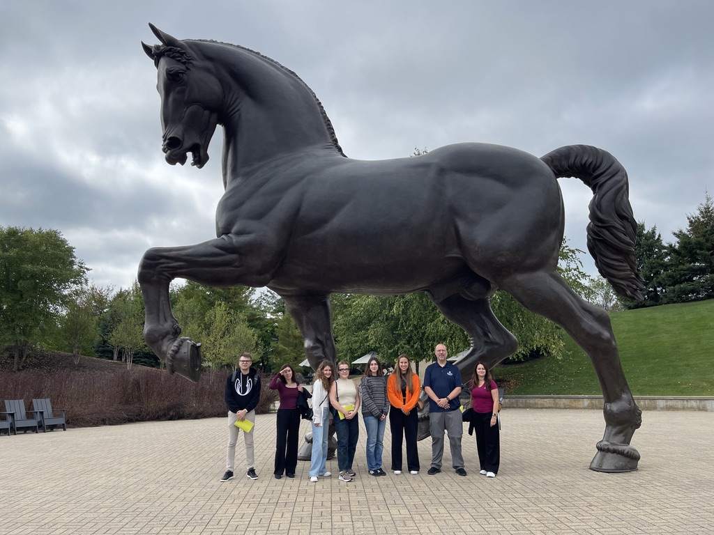Na zdjęciu znajduje się grupa ośmiu osób ustawionych w szeregu na tle monumentalnej, ciemnej rzeźby konia w dynamicznej pozie. Postument z figurą stoi na utwardzonym, jasnym placu, a w tle widoczne są drzewa i zielone wzgórze. Osoby mają na sobie codzienne stroje, w rękach trzymają torby oraz dokumenty. Niebo jest pochmurne, co podkreśla monumentalny charakter rzeźby.