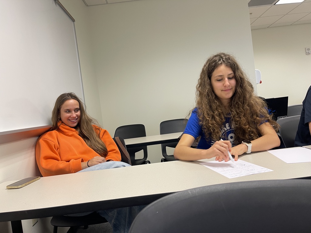 Na zdjęciu widoczne są dwie osoby w sali wykładowej. Osoba po lewej stronie siedzi pod ścianą, ubrana w pomarańczową bluzę i niebieskie jeansy, a jej postawa jest swobodna. Osoba po prawej, w niebieskiej koszulce, intensywnie zapisuje kartkę papieru przy biurku. Tło stanowi jasna ściana z fragmentem tablicy, krzesła ustawione są w rzędach, a wystrój pomieszczenia jest skromny i typowy dla uczelni lub szkoły.