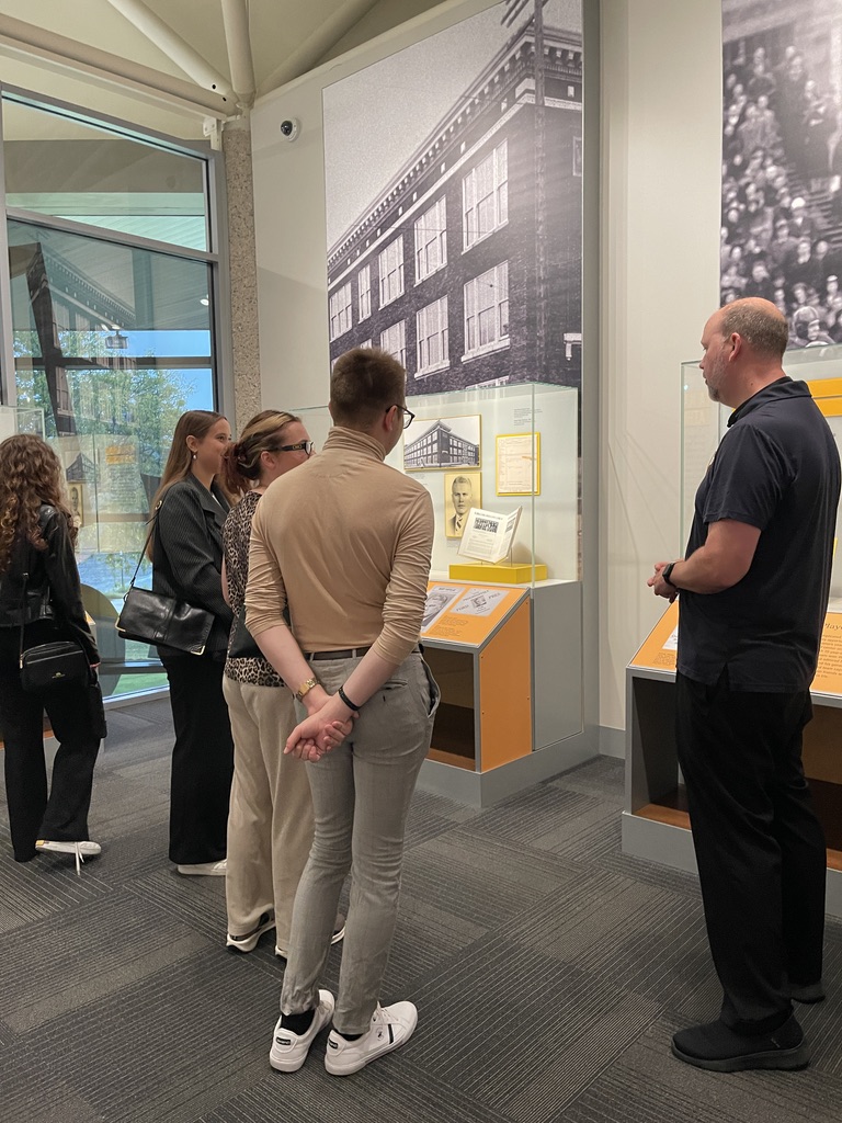 Na zdjęciu znajduje się kilka osób zwiedzających budynek muzeum. Grupa stoi przed witrynami z eksponatami oraz planszami informacyjnymi, poświęconymi architekturze oraz wydarzeniom z przeszłości. Osoby w centrum kadru są zwrócone twarzami do gablot, rozmawiając lub czytając opisy. W tle widoczne są duże czarno-białe zdjęcia budynków i tłumu ludzi, a po lewej stronie znajduje się przeszklona ściana, przez którą wpada naturalne światło. Pomieszczenie urządzone jest w nowoczesnym stylu, z szarymi wykładzinami i przeszklonymi elementami.