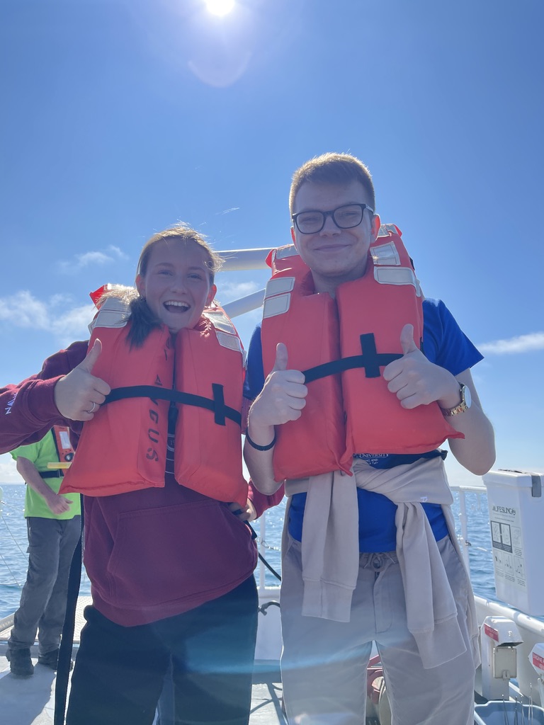 Na zdjęciu znajdują się dwie osoby stojące na pokładzie łodzi w słoneczny dzień. Oboje mają na sobie pomarańczowe kamizelki ratunkowe i wykonują gest „kciuk w górę” w kierunku aparatu. Osoba po lewej ubrana jest w bordową bluzę, a osoba po prawej w niebieską koszulkę oraz jasne spodnie z przewiązaną bluzą w pasie. W tle widoczni są pozostali uczestnicy rejsu, fragment morza lub jeziora oraz jasne niebo z wyraźnym słońcem umieszczonym w górnej części kadru.
