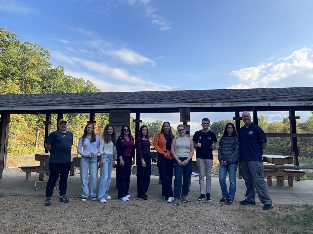 Na zdjęciu znajduje się grupa dziesięciu osób stojących w rzędzie przed zadaszonym pawilonem piknikowym, w otoczeniu zieleni i drzew. Osoby są ubrane w casualowe stroje – jeansy, swetry, t-shirty i bluzy, jedna kobieta wyróżnia się pomarańczową narzutką. W tle widoczne są drewniane ławki i stoły pod zadaszeniem, tabliczka z regulaminem oraz piękne, lekko jesienne drzewa w promieniach popołudniowego słońca. Grupa pozuje na pamiątkowym zdjęciu, a atmosfera jest swobodna i przyjazna.