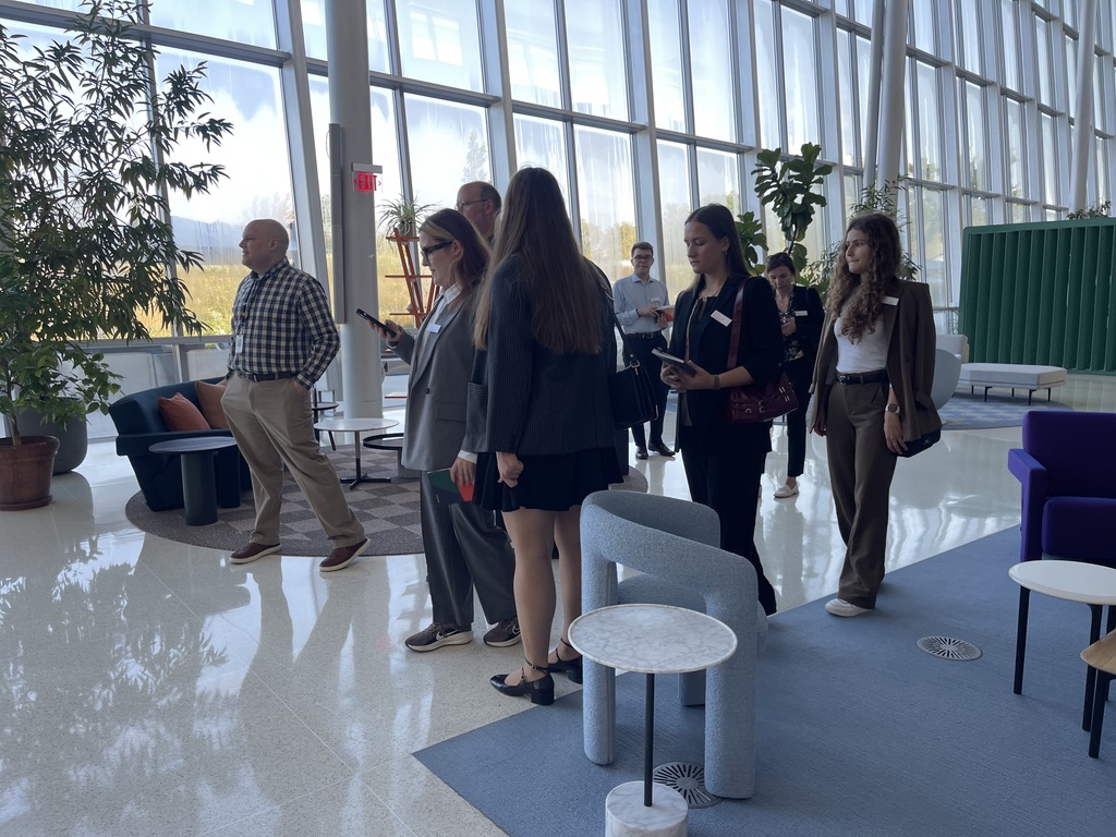 Na zdjęciu widoczna jest grupa osób zwiedzających nowoczesne, przestronne wnętrze o wysokich, przeszklonych ścianach – budynki firmy HAWORTH. Uczestnicy są elegancko ubrani – wiele osób ma na sobie marynarki i garsonki, a niektórzy trzymają notesy lub telefony. W tle znajdują się duże doniczkowe rośliny, designerskie fotele oraz strefa wypoczynkowa z poduszkami i stolikami. Naturalne światło wpada do wnętrza przez wysokie okna, nadając przestrzeni jasny i przyjazny charakter. Cała scena ma profesjonalny, konferencyjny klimat.