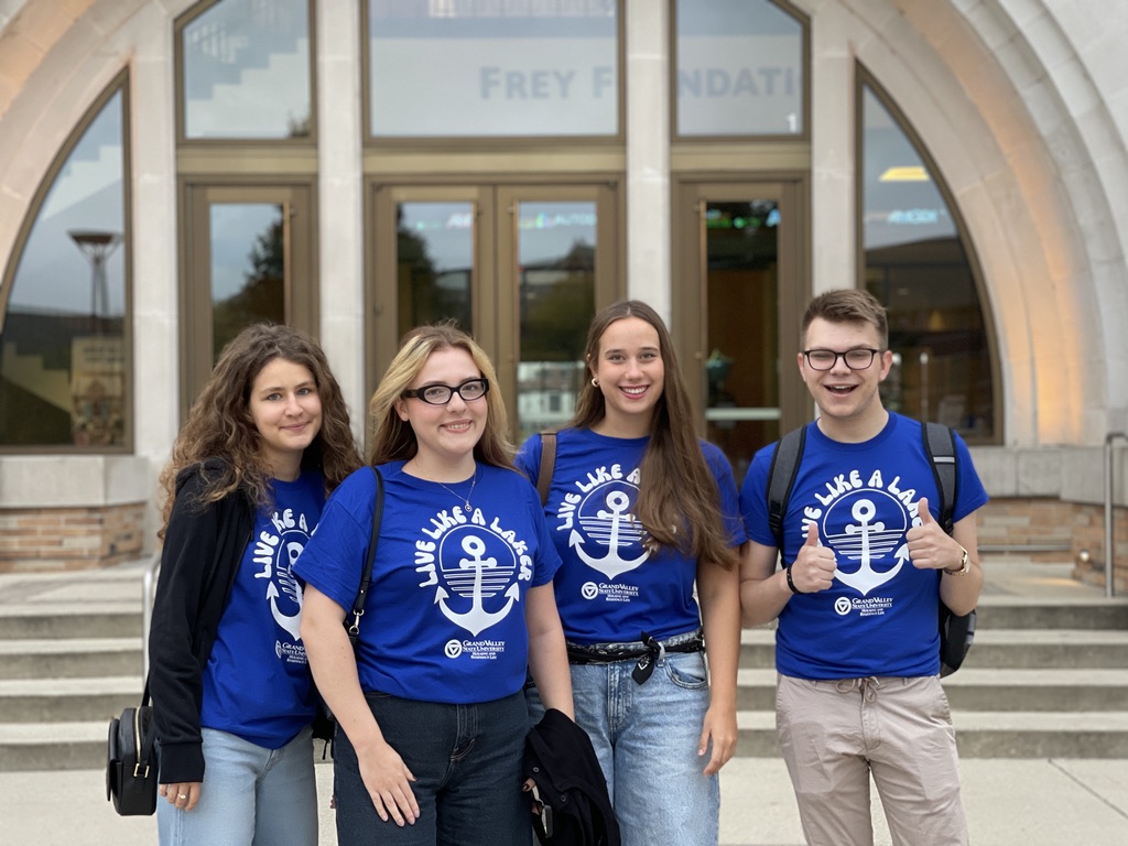 Na zdjęciu znajduje się czworo młodych ludzi stojących razem przed wejściem do budynku, na którym widoczny jest napis „Frey Foundation”. Wszyscy mają na sobie niebieskie koszulki z nadrukiem „Live Like a Lake” i symbolem kotwicy, a dwoje z nich ma na plecach czarne plecaki. Osoby uśmiechają się i stoją blisko siebie, a jedna z nich pokazuje gest „kciuk w górę” obydwiema rękami. Tło stanowią charakterystyczne, półokrągłe łuki budynku oraz przeszklone drzwi wejściowe. Atmosfera zdjęcia jest radosna i integracyjna.