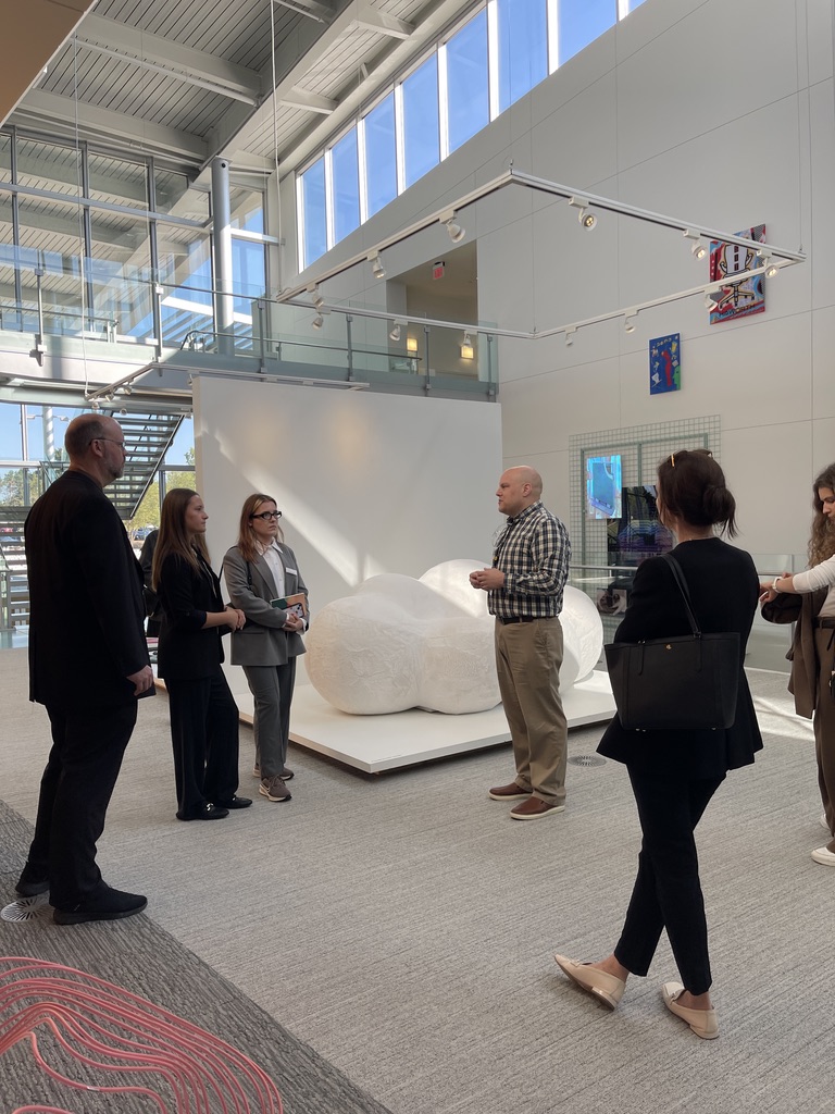 Na zdjęciu znajduje się grupa siedmiu osób, rozmawiających i zwiedzających nowoczesne wnętrze firmy HAROWTH. W centrum kadru widoczna jest biała abstrakcyjna rzeźba umieszczona na podestach. Osoby są elegancko ubrane – mężczyzna w kratkowanej koszuli prowadzi rozmowę, a pozostali mają na sobie garnitury, marynarki lub sukienki. W tle widać przeszklone ściany i piętro budynku z balustradą. Całość wnętrza jest jasna, przestronna i nowoczesna, a atmosfera sprzyja profesjonalnym spotkaniom i inspiracjom artystycznym.