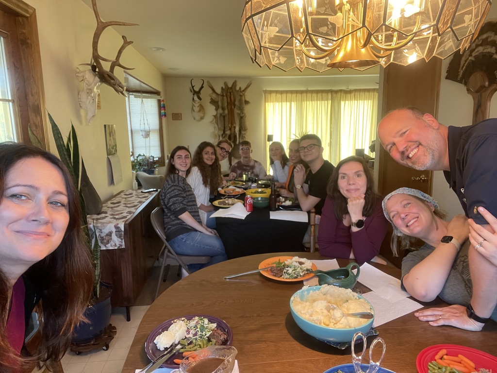 Na zdjęciu widoczna jest grupa osób zgromadzonych przy dwóch stołach w przytulnym, domowym wnętrzu. Na pierwszym planie uśmiechnięta kobieta robi selfie, a na stole widać porcje jedzenia, sałatkę, ziemniaki i napoje. Dalej, przy większym stole, kilkoro uczestników siedzi blisko siebie, rozmawia i spożywa posiłek – atmosfera jest rodzinna i swobodna. W tle na ścianach widać trofea myśliwskie, rzeźby zwierzęcych głów oraz rośliny doniczkowe. Pomieszczenie jest jasne i ciepłe dzięki wpadającemu światłu oraz dużej lampie nad stołem.