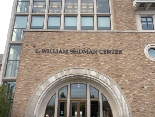 Na zdjęciu znajduje się grupa sześciu osób stojących przed wejściem do budynku L. William Seidman Center. Budynek wyróżnia się nowoczesną, wysoką fasadą wykonaną z cegły i dużymi oknami. Nad wejściem widoczny jest łuk oraz napis z nazwą obiektu. Osoby stoją na szerokim chodniku prowadzącym do schodów i głównych drzwi. W tle widać fragment zieleni oraz niebo z lekkim zachmurzeniem.