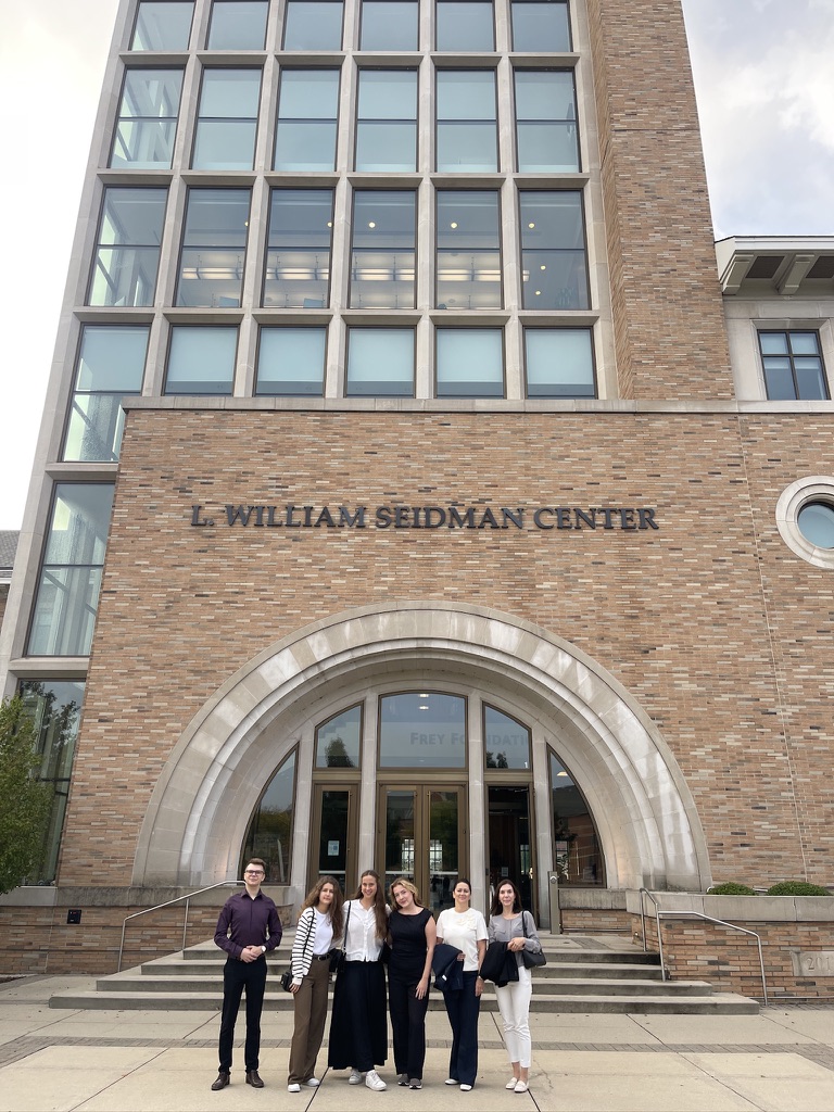 Na zdjęciu znajduje się grupa sześciu osób stojących przed wejściem do budynku L. William Seidman Center. Budynek wyróżnia się nowoczesną, wysoką fasadą wykonaną z cegły i dużymi oknami. Nad wejściem widoczny jest łuk oraz napis z nazwą obiektu. Osoby stoją na szerokim chodniku prowadzącym do schodów i głównych drzwi. W tle widać fragment zieleni oraz niebo z lekkim zachmurzeniem.