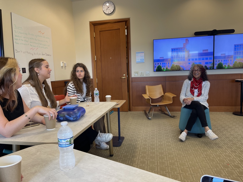 Na zdjęciu znajduje się grupa czterech osób siedzących przy dwóch stołach ustawionych w literę L po lewej stronie kadru. Każda z tych osób trzyma w ręku papierowy kubek, a na stole stoją dodatkowo butelka wody oraz pojemnik śniadaniowy. Po prawej stronie, naprzeciwko grupy, siedzi kobieta z ciemnymi włosami - DIANA LAWSON – Dean, Seidman College of Business, ubrana w jasny sweter i czerwony szalik. W tle, na ścianie, widoczne są dwa połączone monitory z widokiem budynku oraz zamknięte drzwi z zegarem powieszonym nad nimi. Pokój jest jasny, z wykładziną w stonowanych kolorach.